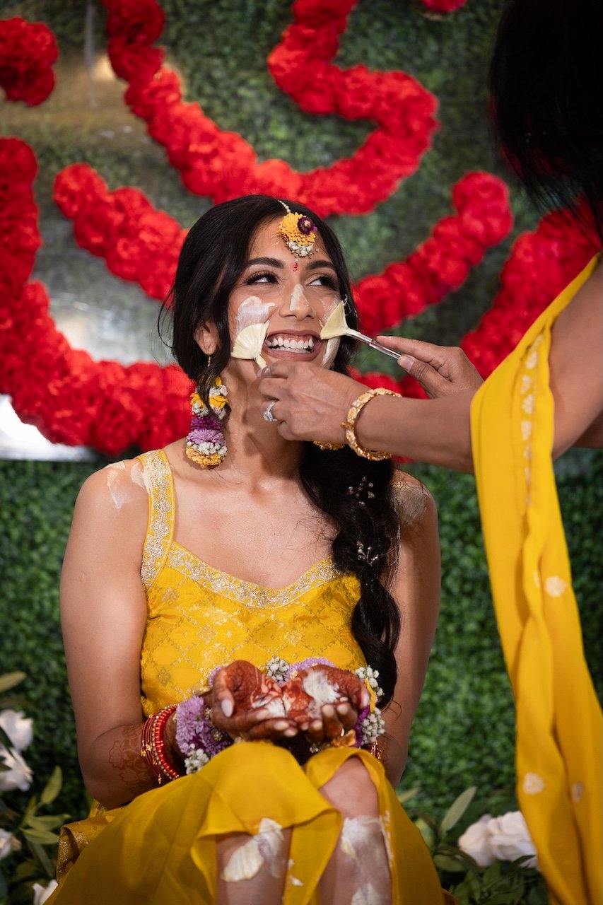 Bride in yellow traditional wedding dress having turmeric and other ritual powders applied to her face during a pre-wedding ceremony, decorated with flowers and marigolds.
