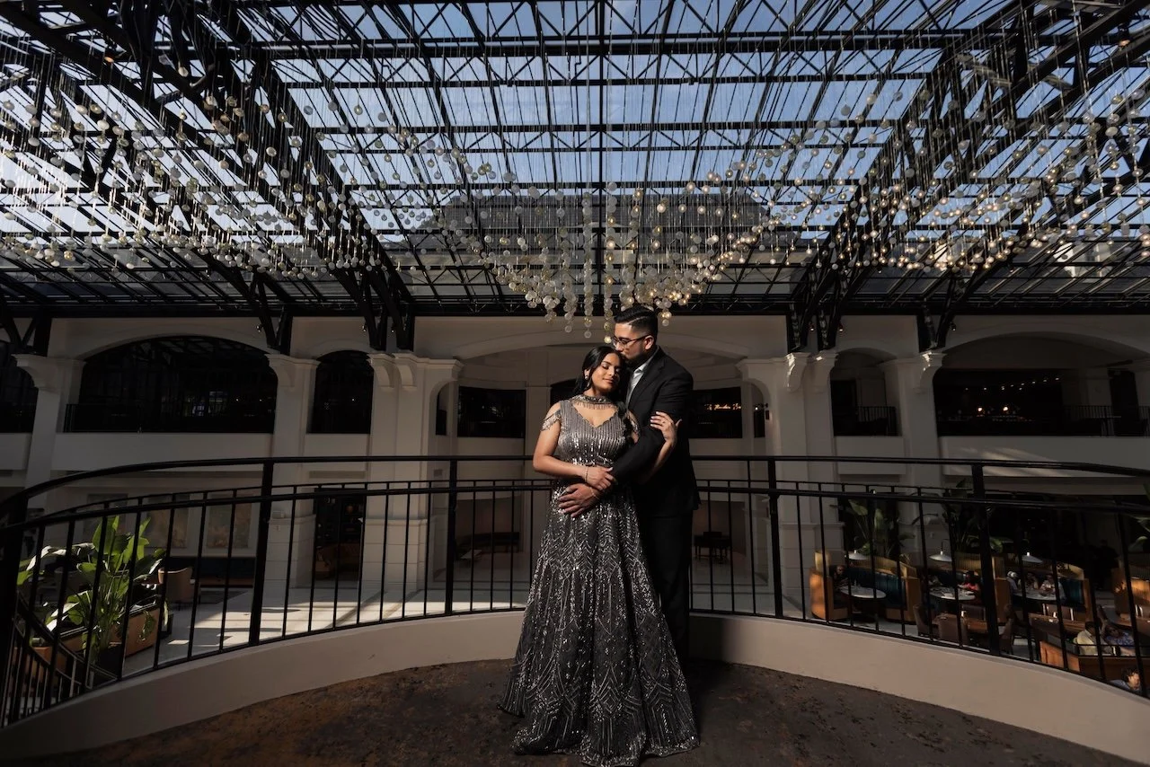 A man and woman in formal attire standing close together on a balcony inside a building with a glass ceiling and hanging lights, possibly at a wedding or event.