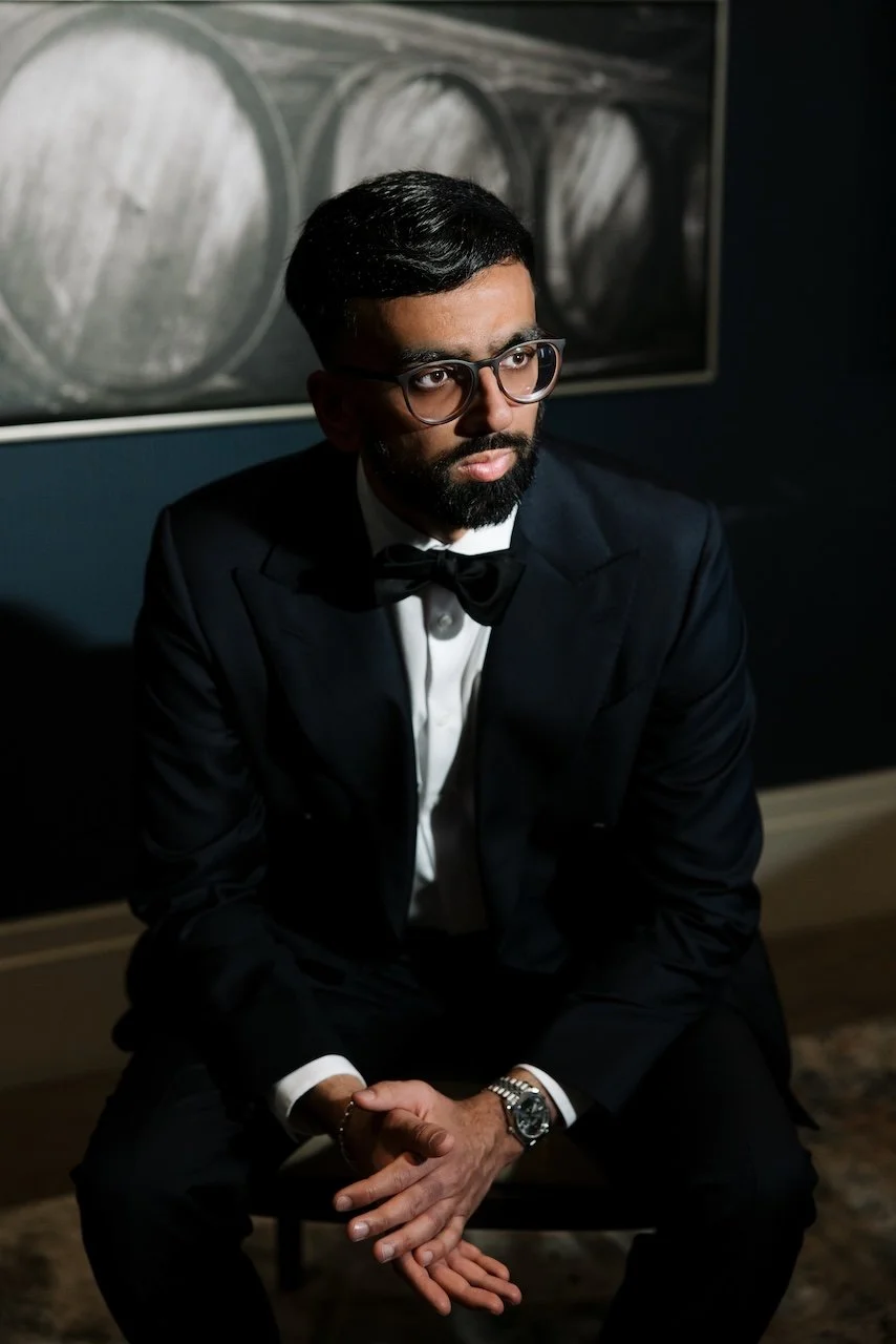A man dressed in a black tuxedo with a bow tie, sitting with hands clasped, wearing glasses, with a beard and mustache, in a dimly lit room with artwork in the background.