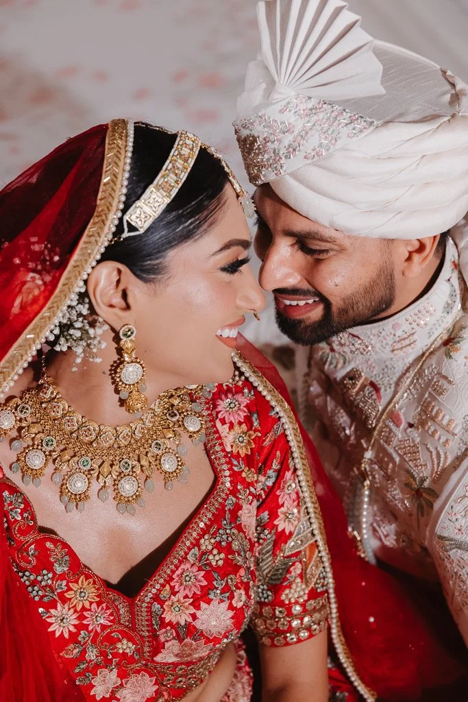 A bride in a red and gold embroidered traditional Indian wedding outfit with jewelry, and a groom in white and silver attire with a turban, are smiling and close together during their wedding ceremony.