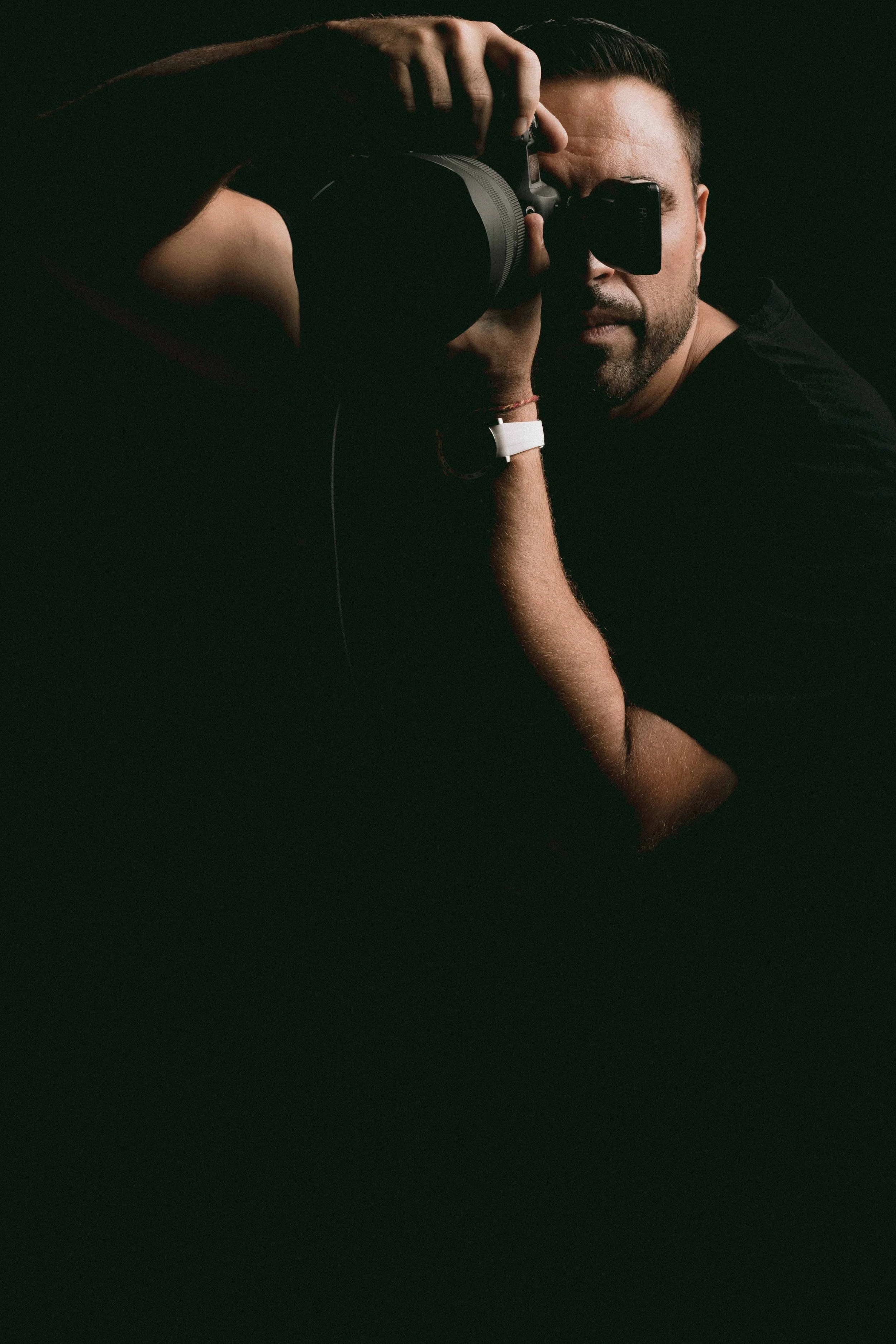A man with dark hair and beard holding a camera up to his face as he takes a picture, wearing a black shirt and a white watch, against a dark background.