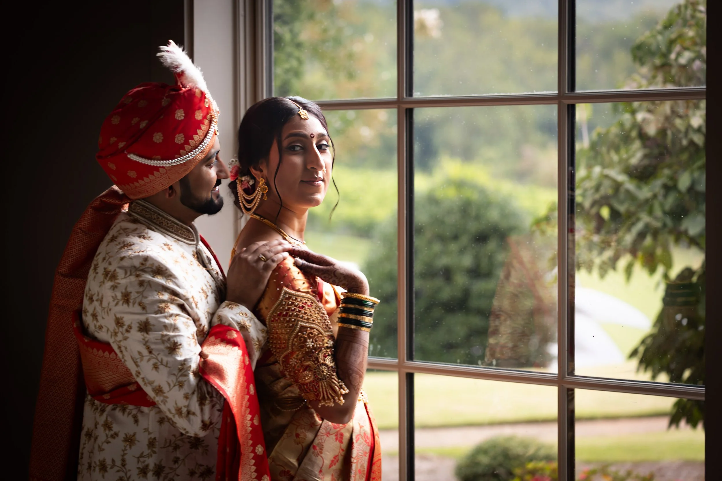 A newlywed Indian couple standing by a window, with both dressed in traditional wedding attire. The groom wears a cream-colored sherwani with red accents and a red turban with white and gold details, while the bride wears a red and gold saree with el