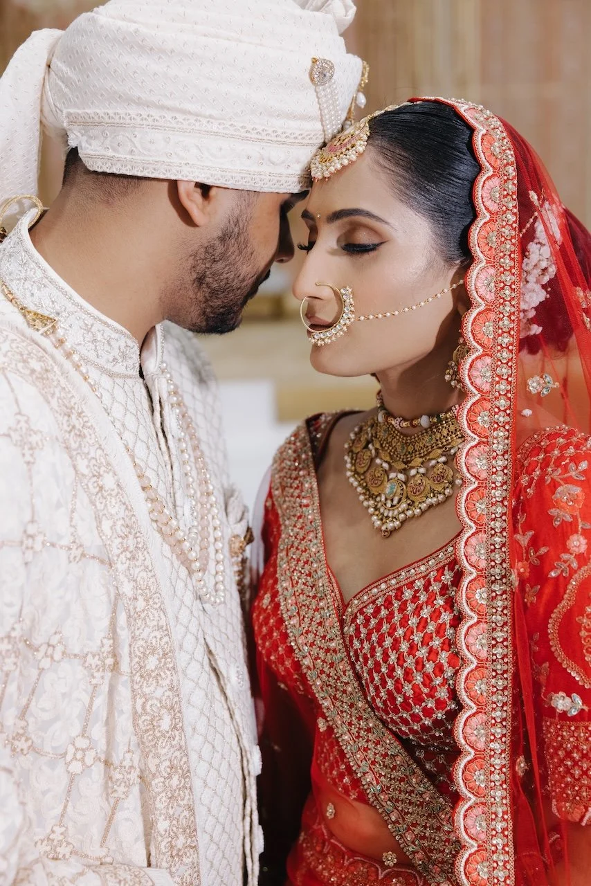 A couple dressed in traditional Indian wedding attire, with their foreheads touching in a close, intimate pose.