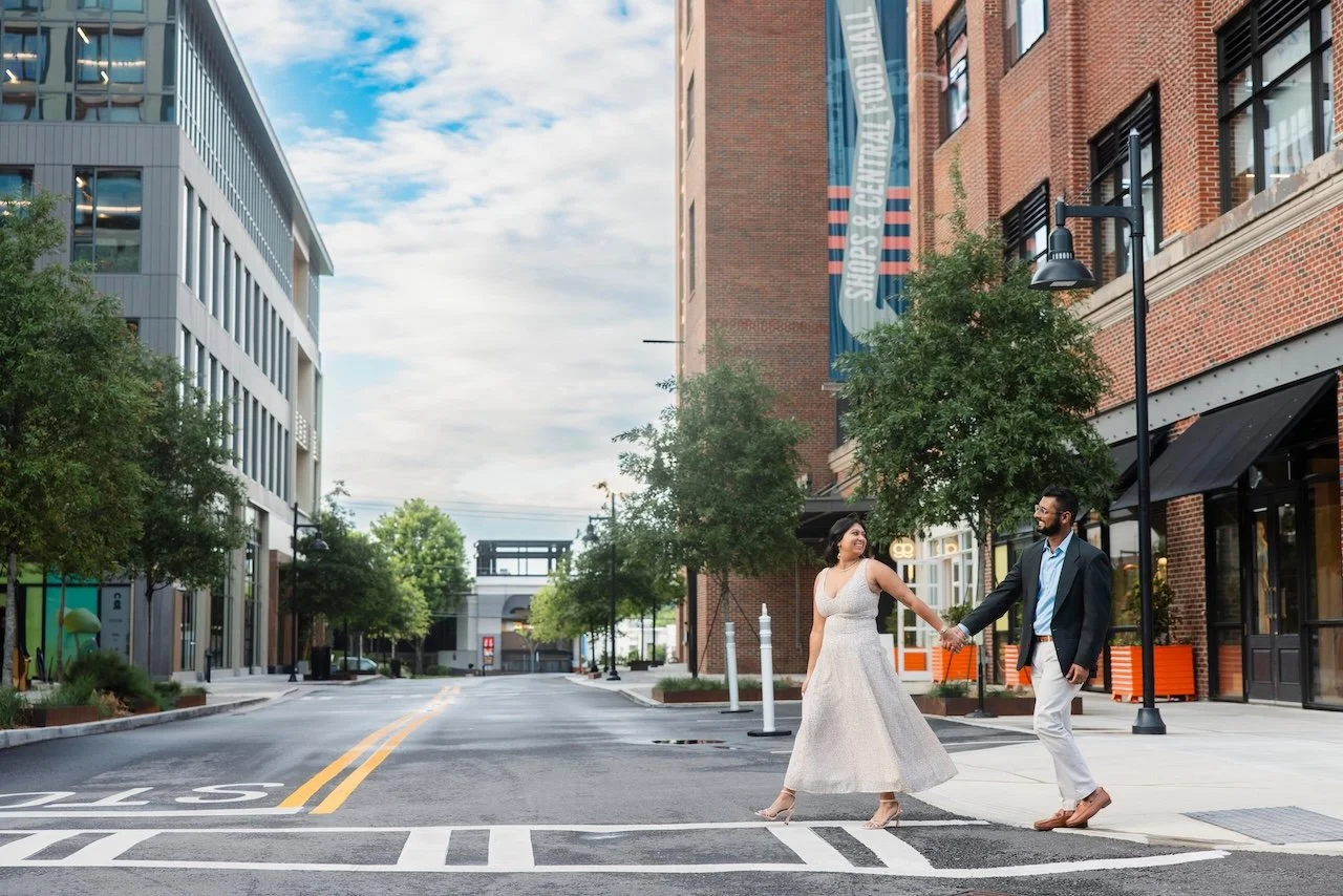 A woman and a man holding hands and walking across an empty city street in an urban area with modern and brick buildings, trees, and streetlights, under a partly cloudy sky.