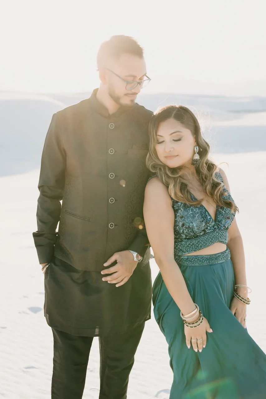 A man and woman standing together on a sandy beach during sunset, dressed in formal attire with the man wearing a black sherwani and the woman in a blue traditional dress with jewelry.