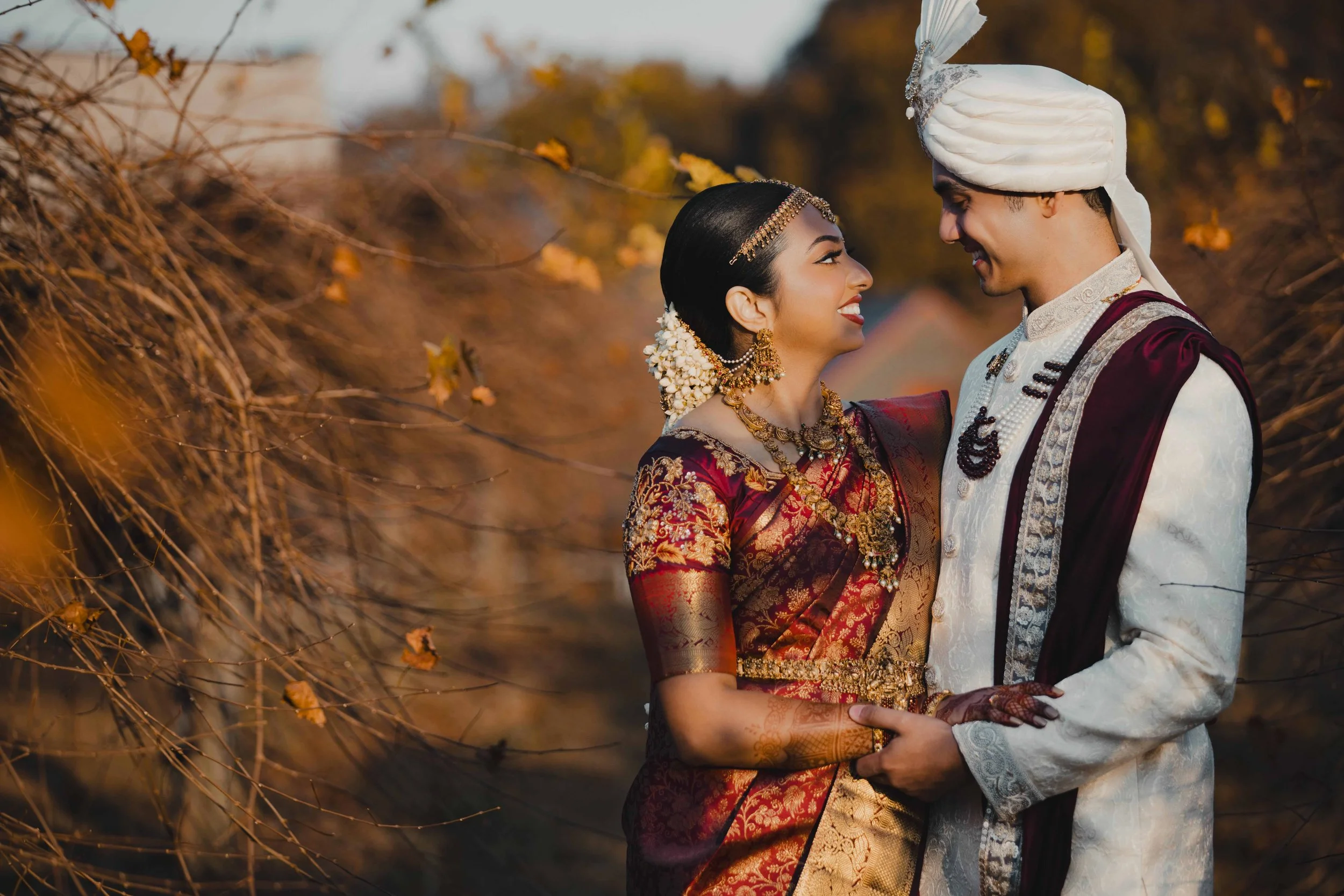A bride and groom in traditional Indian wedding attire outdoors, smiling and looking at each other, surrounded by autumn foliage.