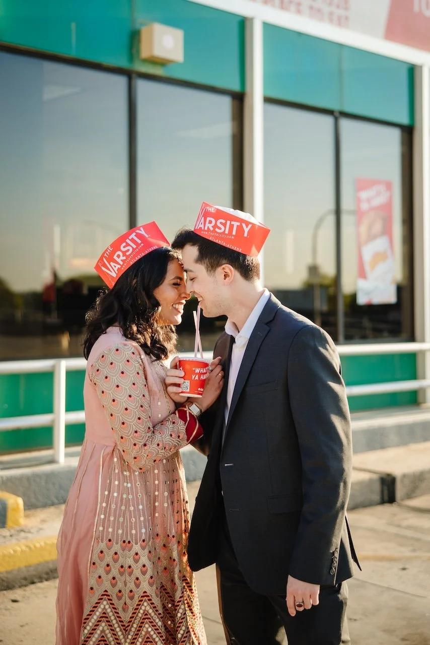 A romantic couple, a woman in a patterned pink dress and a man in a dark suit, are standing outside a building, leaning close with their foreheads touching. They are wearing red paper hats and holding a red cup with a straw between them, smiling happ