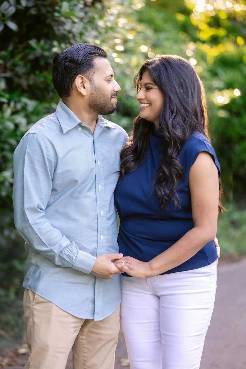 A couple holding hands and smiling at each other outdoors during sunset, surrounded by greenery.