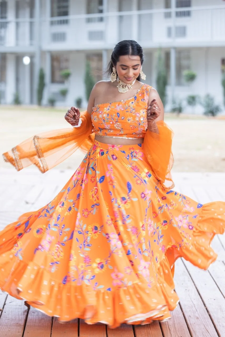 A woman dressed in an orange floral traditional Indian outfit spinning outdoors on a wooden deck, with a white building and greenery in the background.