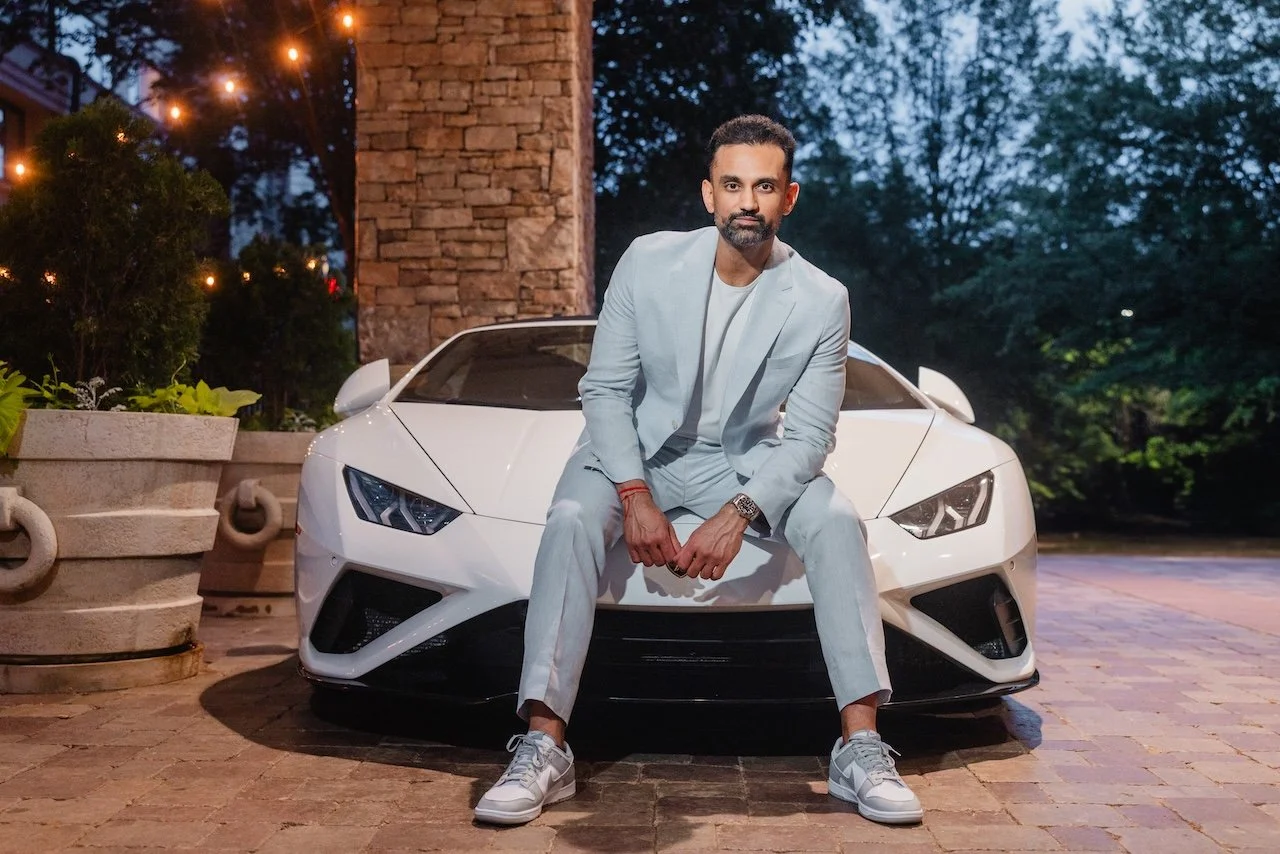 A man wearing a white suit and gray sneakers sitting on the hood of a white luxury sports car, parked on a brick driveway at night with trees and string lights in the background.