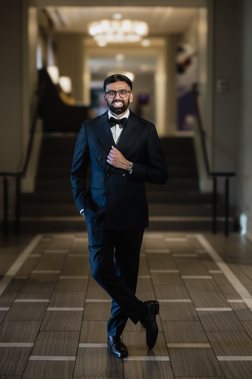 A man in a tuxedo with a bow tie is standing in a fancy corridor, smiling at the camera, with one hand on his chest and the other in his pocket.