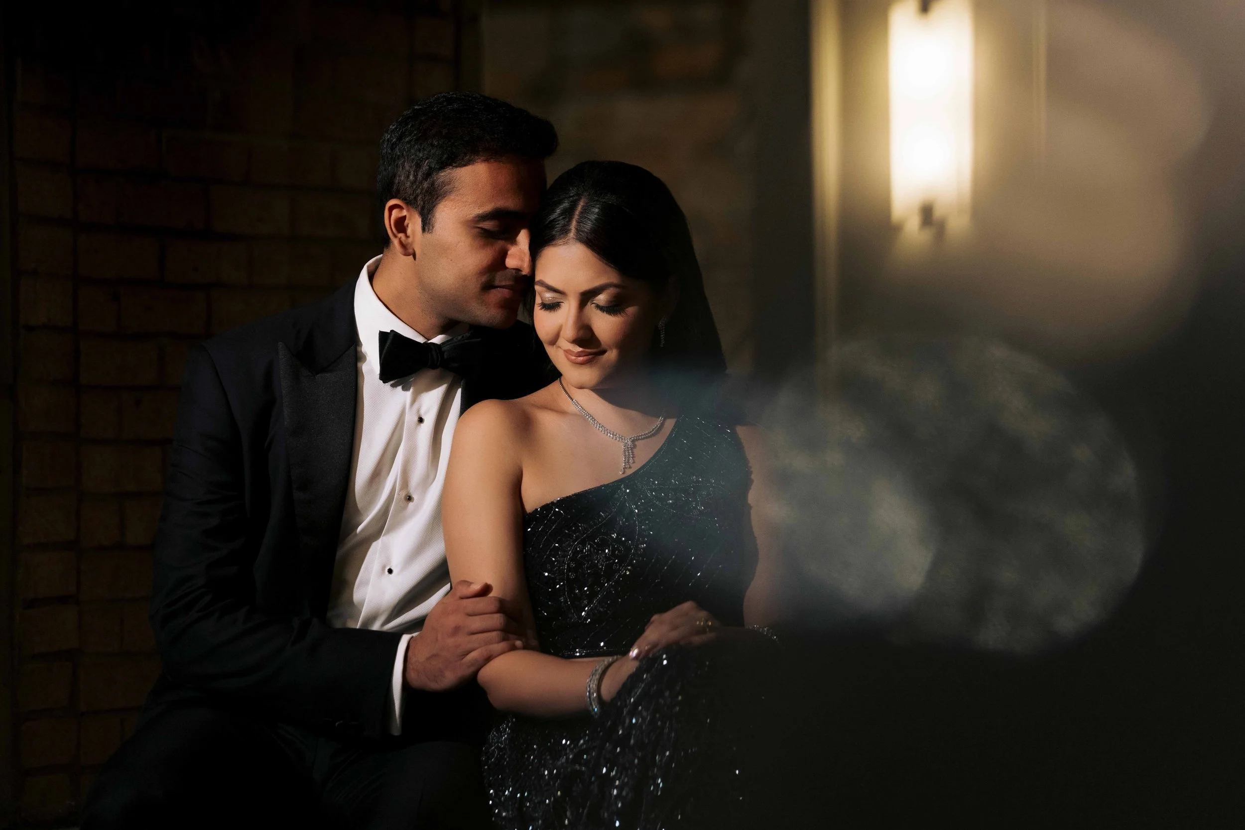 A man and woman dressed in formal attire sitting close together, with the man whispering or nuzzling the woman's ear in a dimly lit setting.