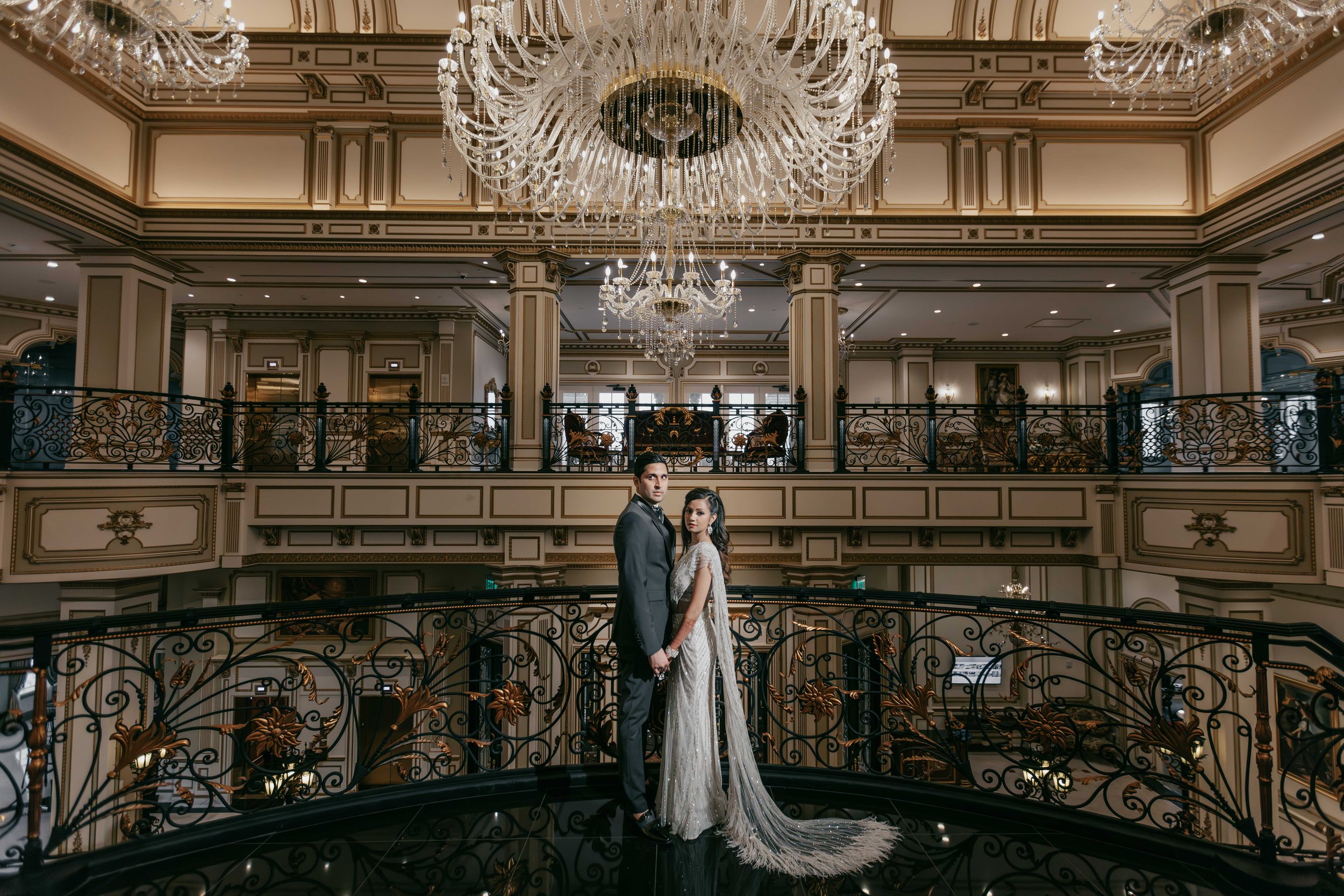 A bride and groom standing hand in hand on a decorative black iron railing in a grand, elegant interior with chandeliers and intricate molding.