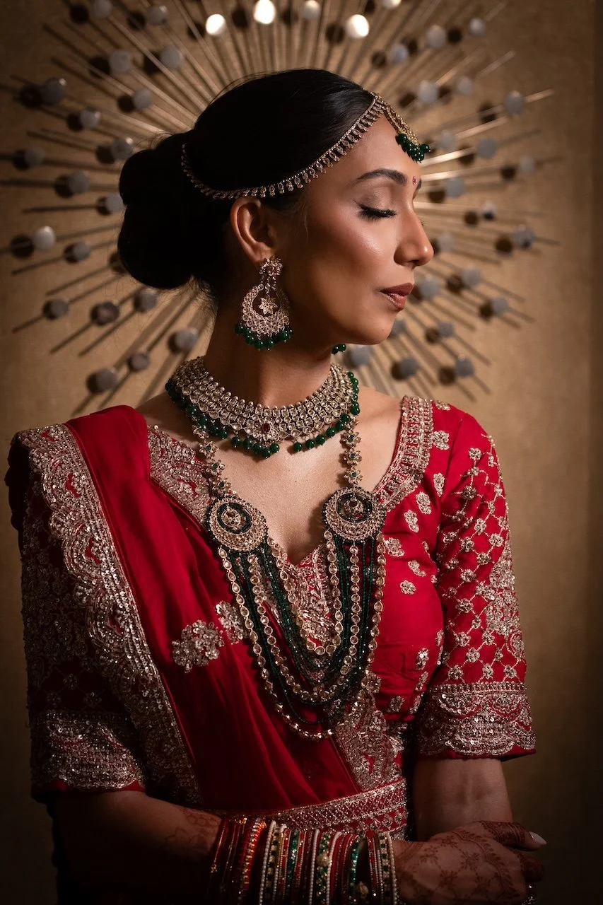 A woman dressed in traditional Indian attire with elaborate jewelry, including earrings, necklaces, and bangles, set against a decorative background with radiating rods and spherical elements.
