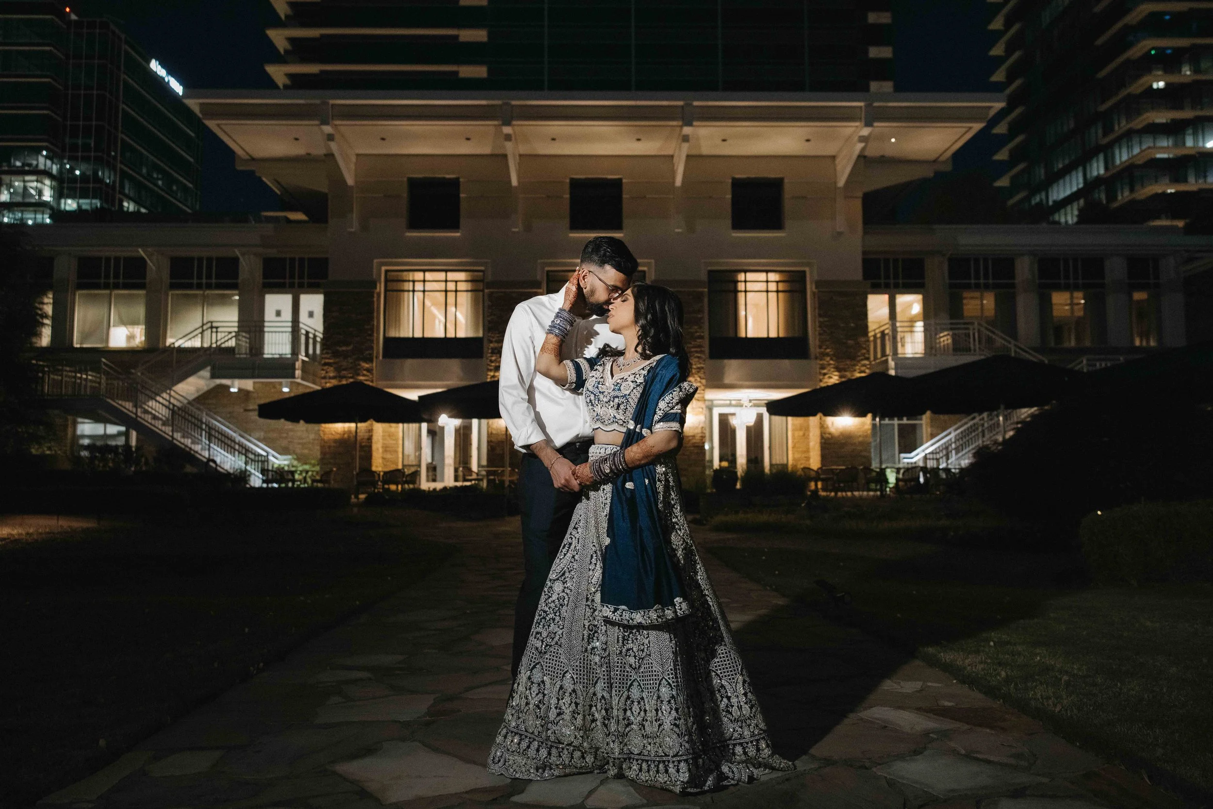 A couple dressed in traditional Indian attire, standing close together on a stone pathway at night, in front of a modern lit building with large windows and balconies, with city buildings in the background.