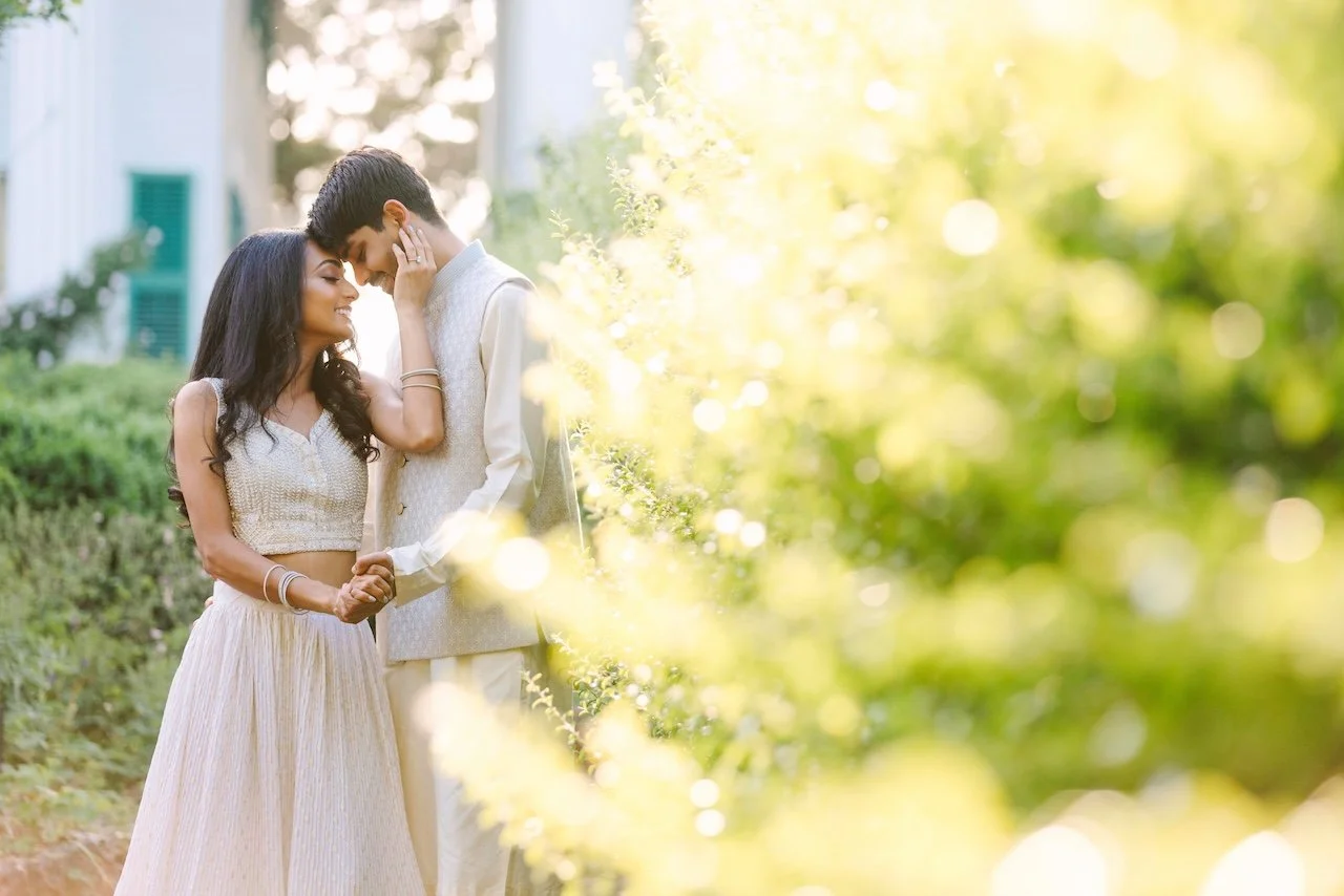 A couple dressed in traditional Indian attire, standing close, touching foreheads, and smiling in an outdoor garden with a house in the background and sunlight filtering through greenery.