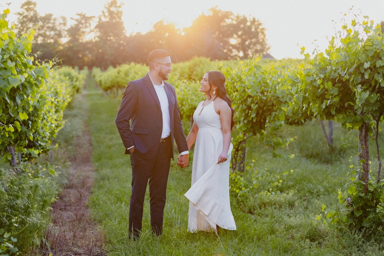 A couple holds hands and looks at each other while standing in a lush green vineyard with rows of grapevines during sunset.