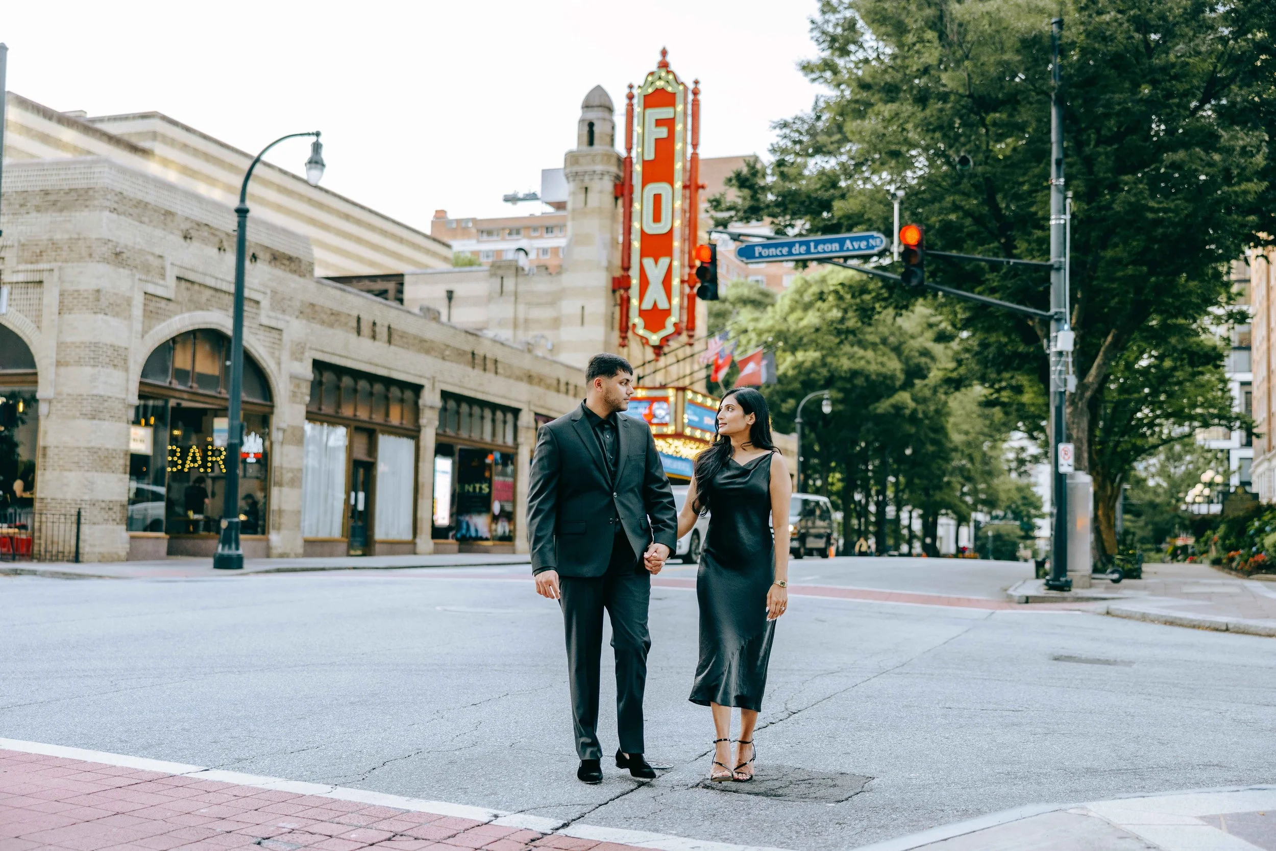 A man and woman in formal attire holding hands and walking across a city street near the Fox Theatre with neon sign and trees.