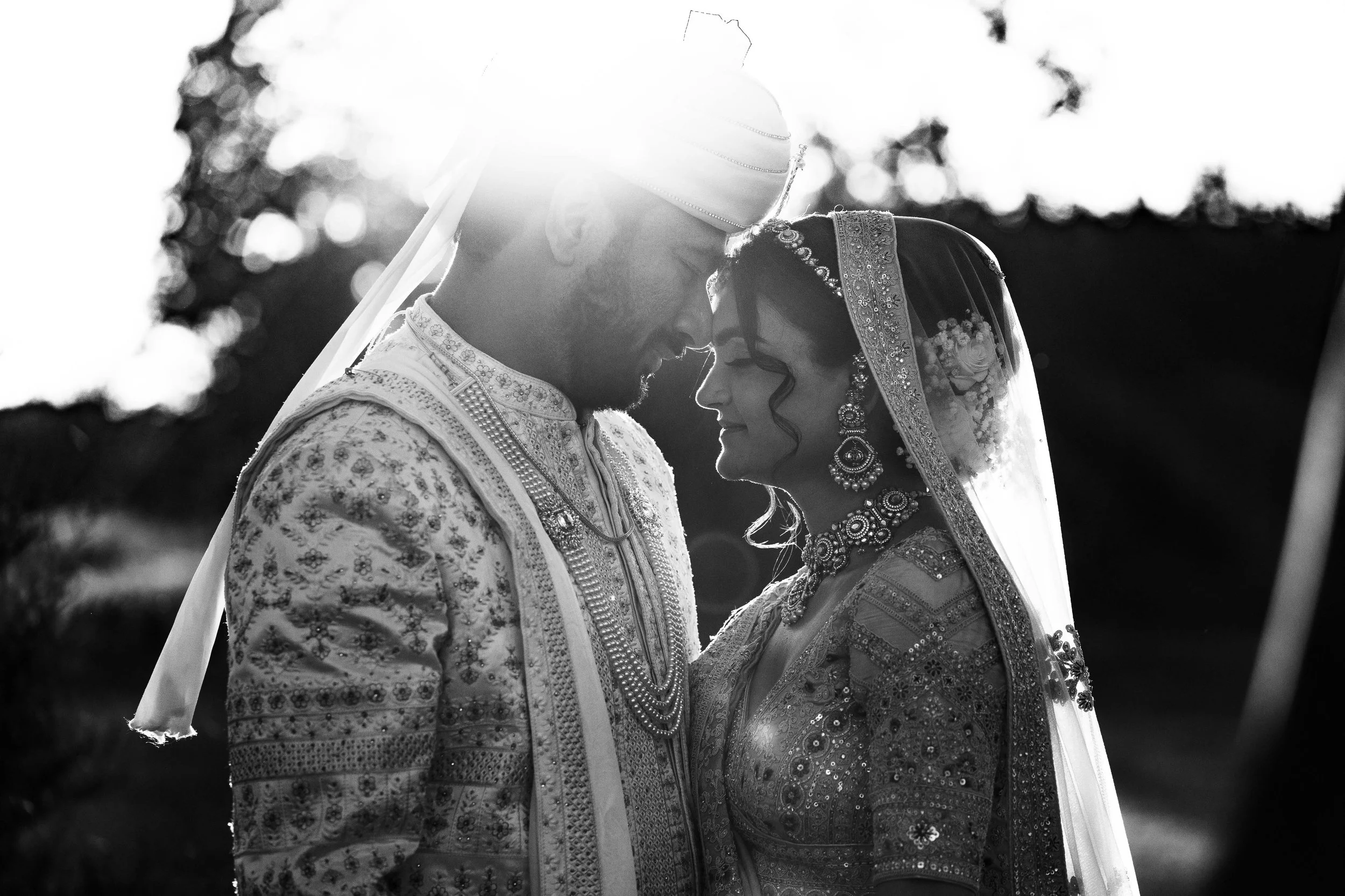 A black and white photo of a bride and groom in traditional Indian wedding attire, standing close with their foreheads touching and eyes closed, backlit by the sun.