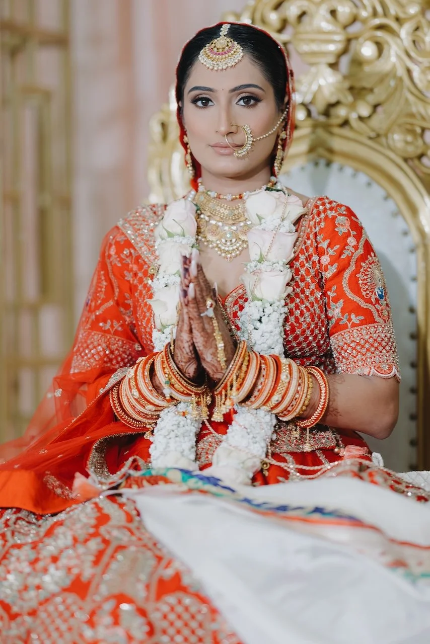 A bride dressed in traditional Indian wedding attire, wearing a red and gold embellished outfit, jewelry, and floral garlands, seated on a golden ornate chair with hands folded in a prayer position.