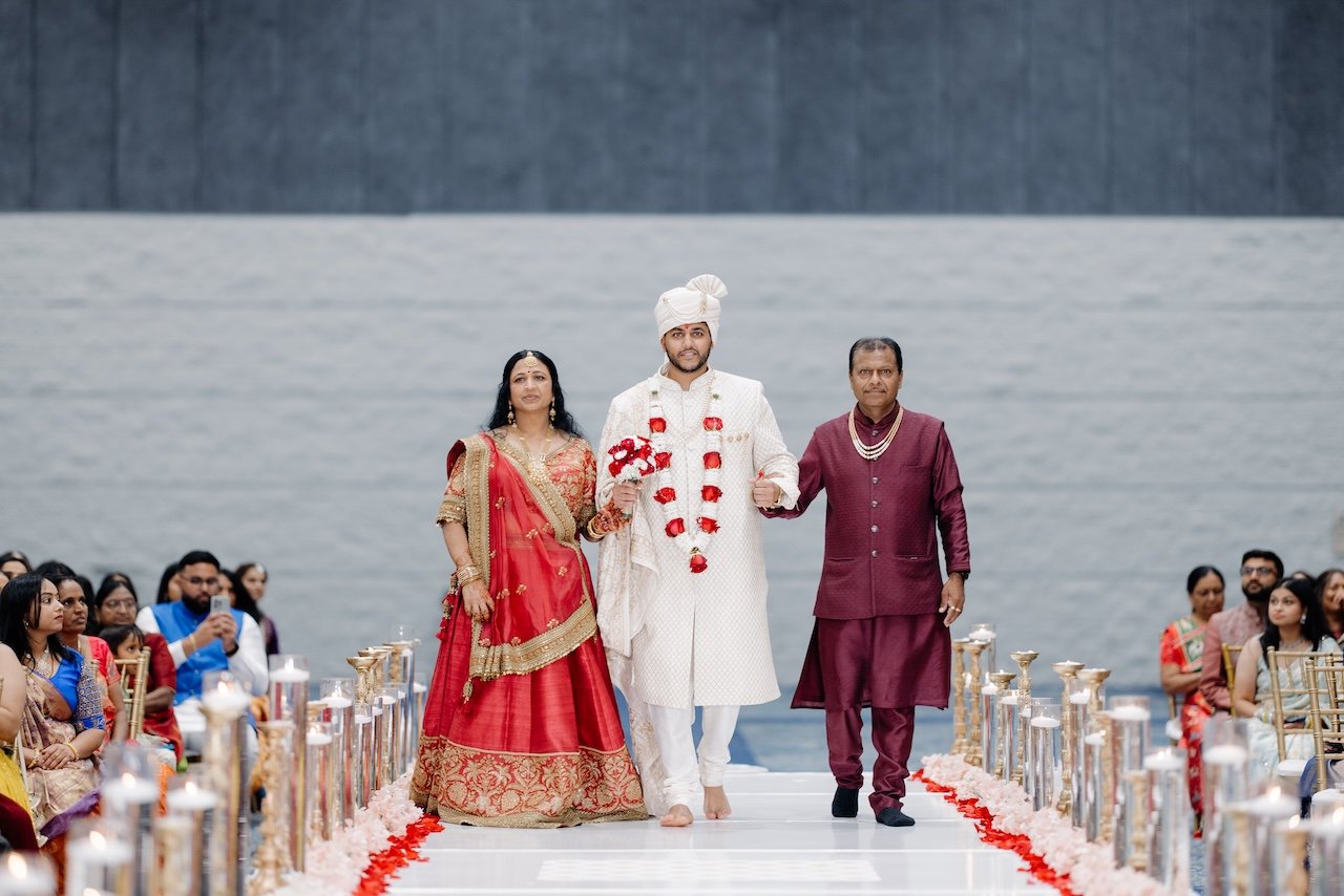 Indian groom in white traditional attire walking down the aisle with his parents at a wedding ceremony, with guests seated on either side, by a body of water.