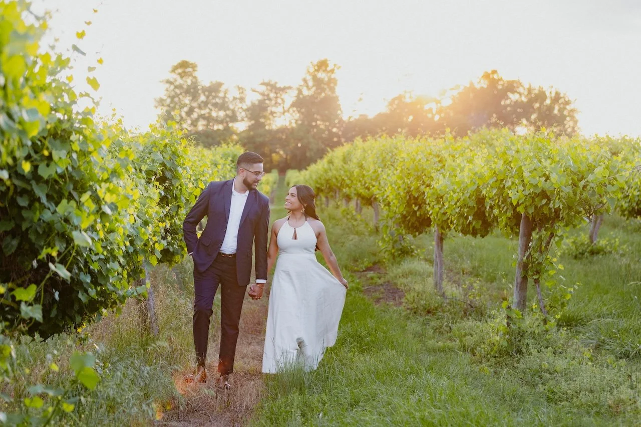 A couple walking hand-in-hand through a vineyard at sunset, with the woman in a white dress and the man in a suit.