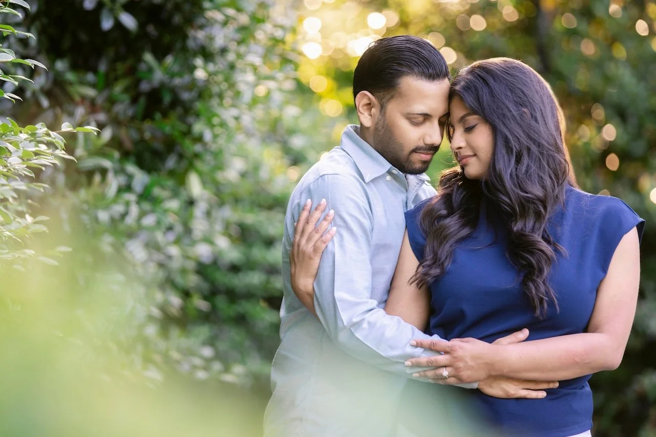 A couple in a garden with green foliage and sunlight, embracing each other with their foreheads touching.