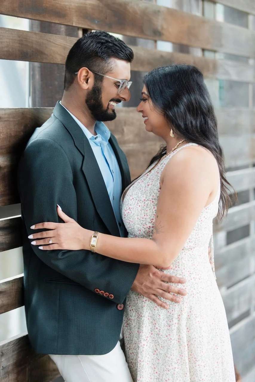 A couple standing close together, smiling with foreheads touching, indoors against a wooden wall.