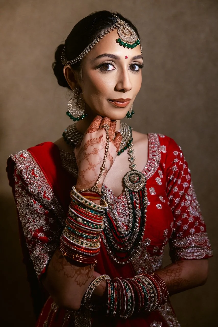 A woman dressed in traditional Indian attire with jewelry, mehndi on her hands, and a red embroidered sari, posing with her hand near her chin.