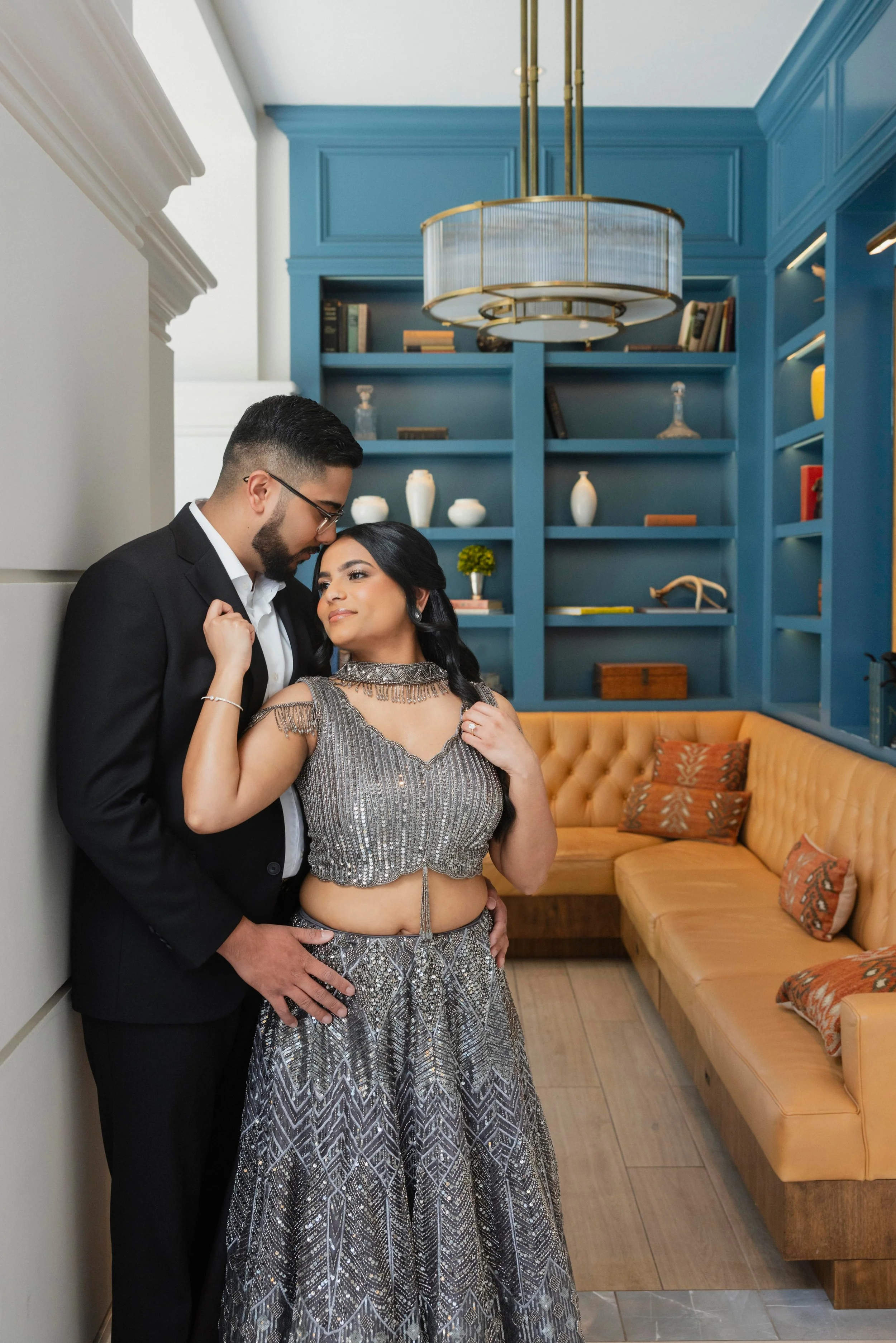 A couple dressed elegantly, with the man in a black suit and glasses and the woman in a shiny silver gown, standing close together in a cozy, stylish living room with a mustard yellow sectional sofa and blue built-in shelves.