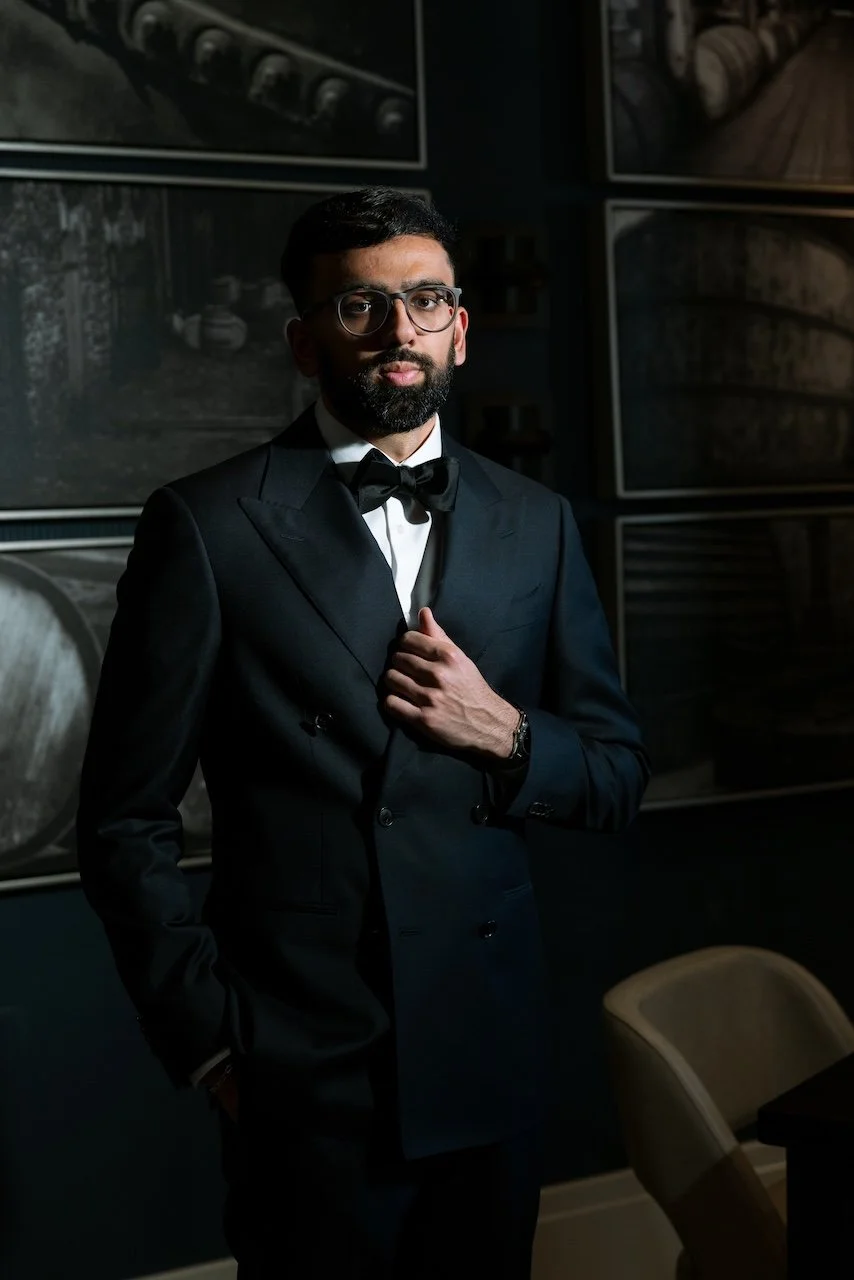 A man wearing glasses and a black tuxedo with a bow tie, standing in a dimly lit room with framed black-and-white photographs on the wall.