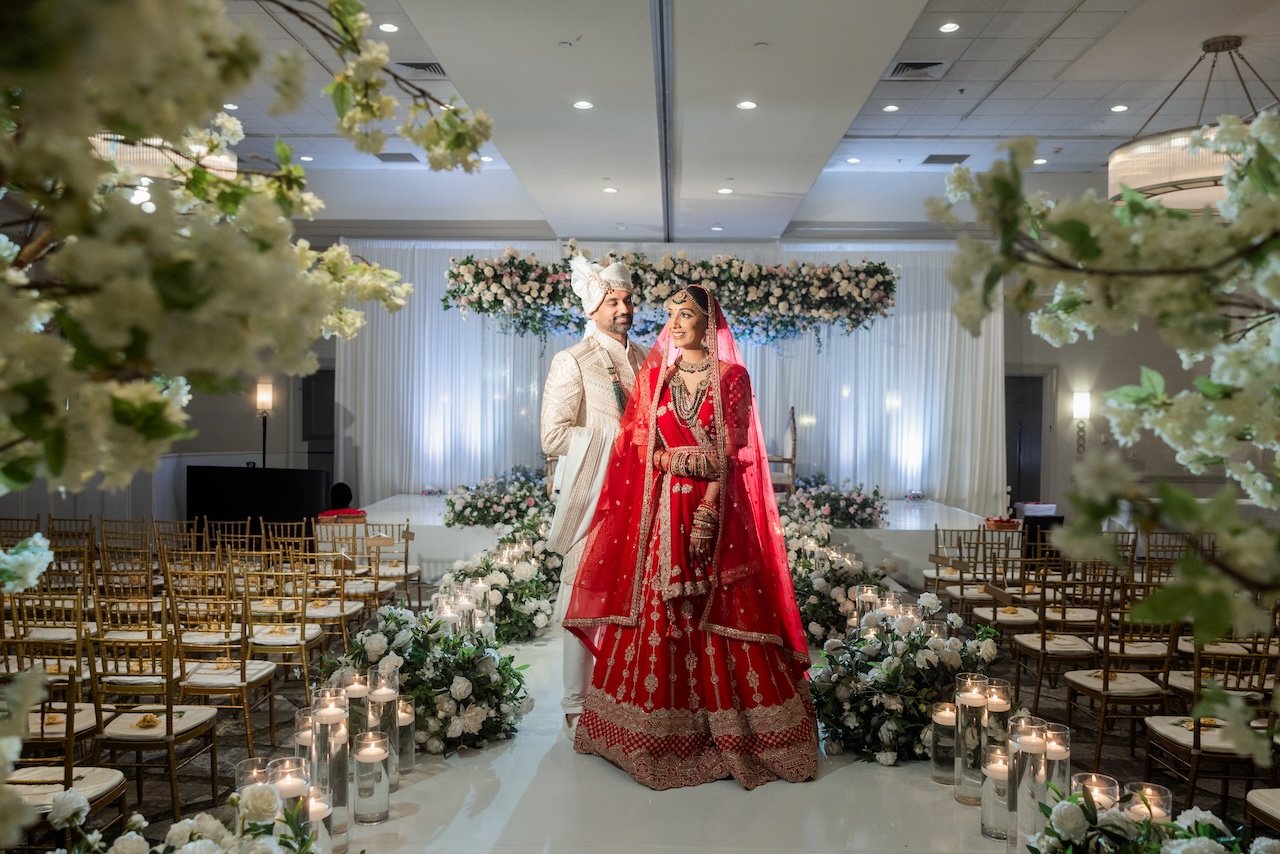 A bride and groom in traditional Indian wedding attire standing in a decorated wedding venue with white flowers, candles, and arranged chairs.