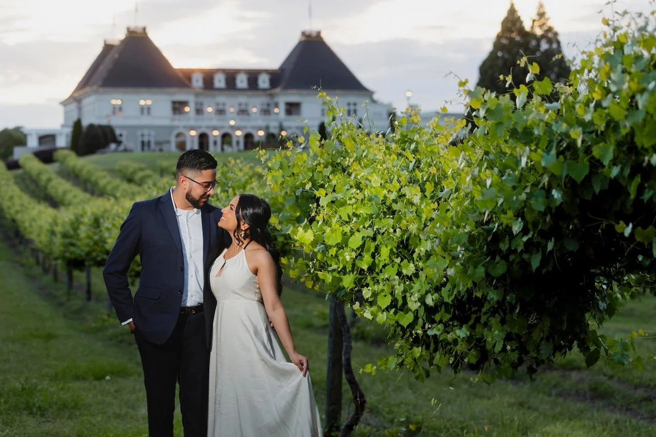 A man in a navy suit and glasses stands close to a woman in a white dress, both smiling and gazing at each other in a vineyard during sunset.