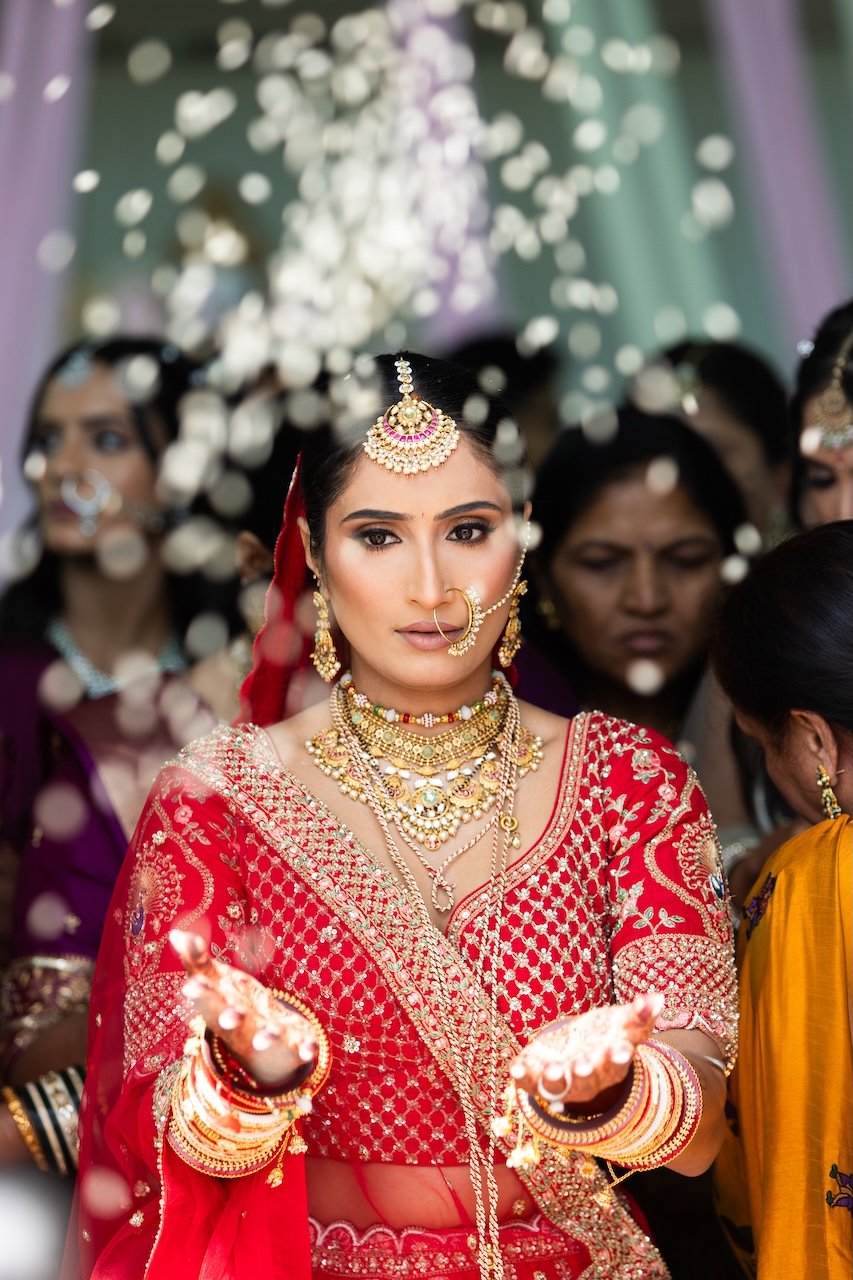A woman dressed in traditional Indian bridal attire, wearing a red embroidered sari with elaborate gold jewelry, including necklaces, earrings, a maang tikka, and a nose ring, participating in a wedding ceremony surrounded by women.