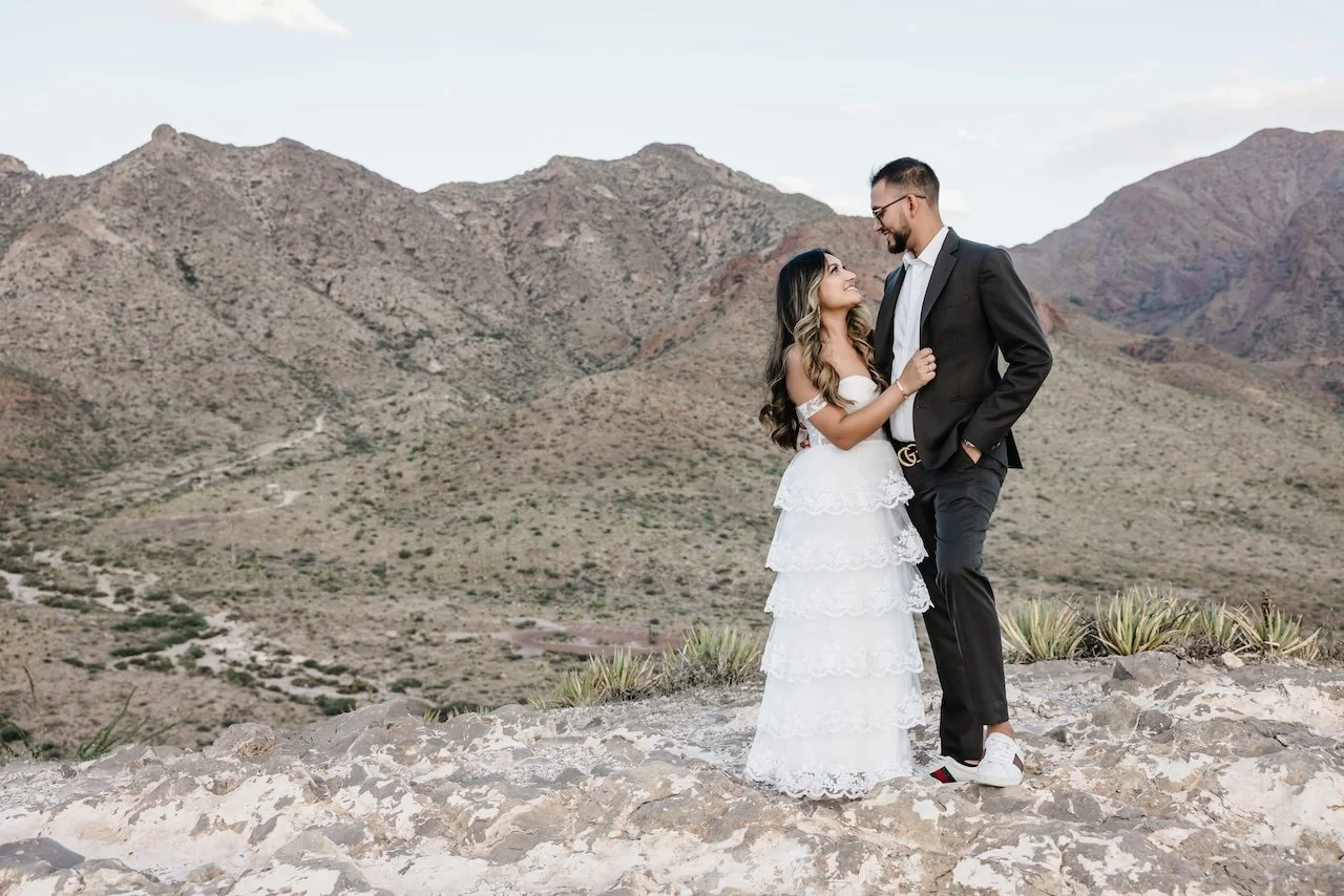 A couple dressed in wedding attire standing on rocky terrain with mountainous desert landscape in the background, sharing an intimate moment.