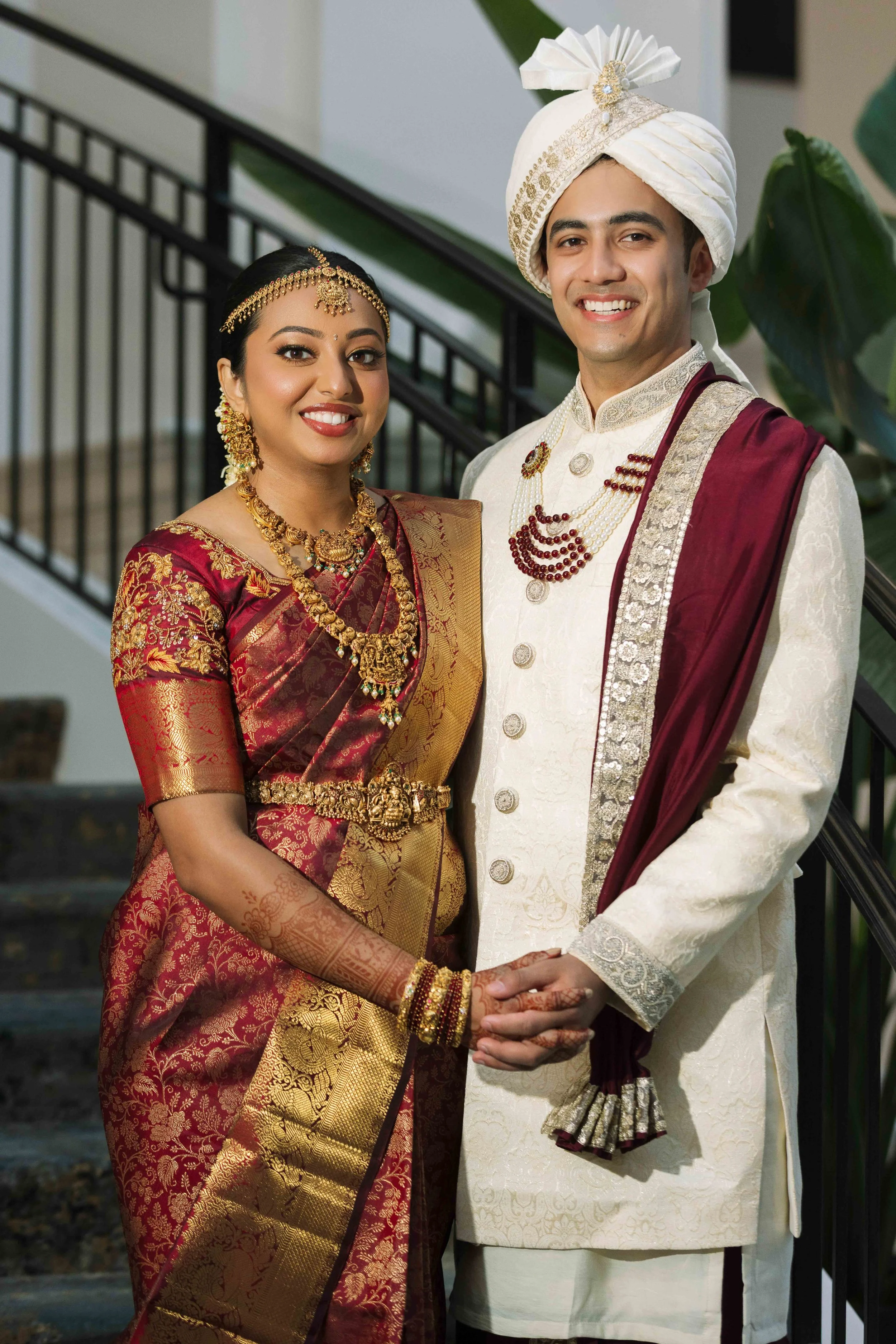 Indian bride and groom in traditional wedding attire, standing hand in hand indoors, smiling.