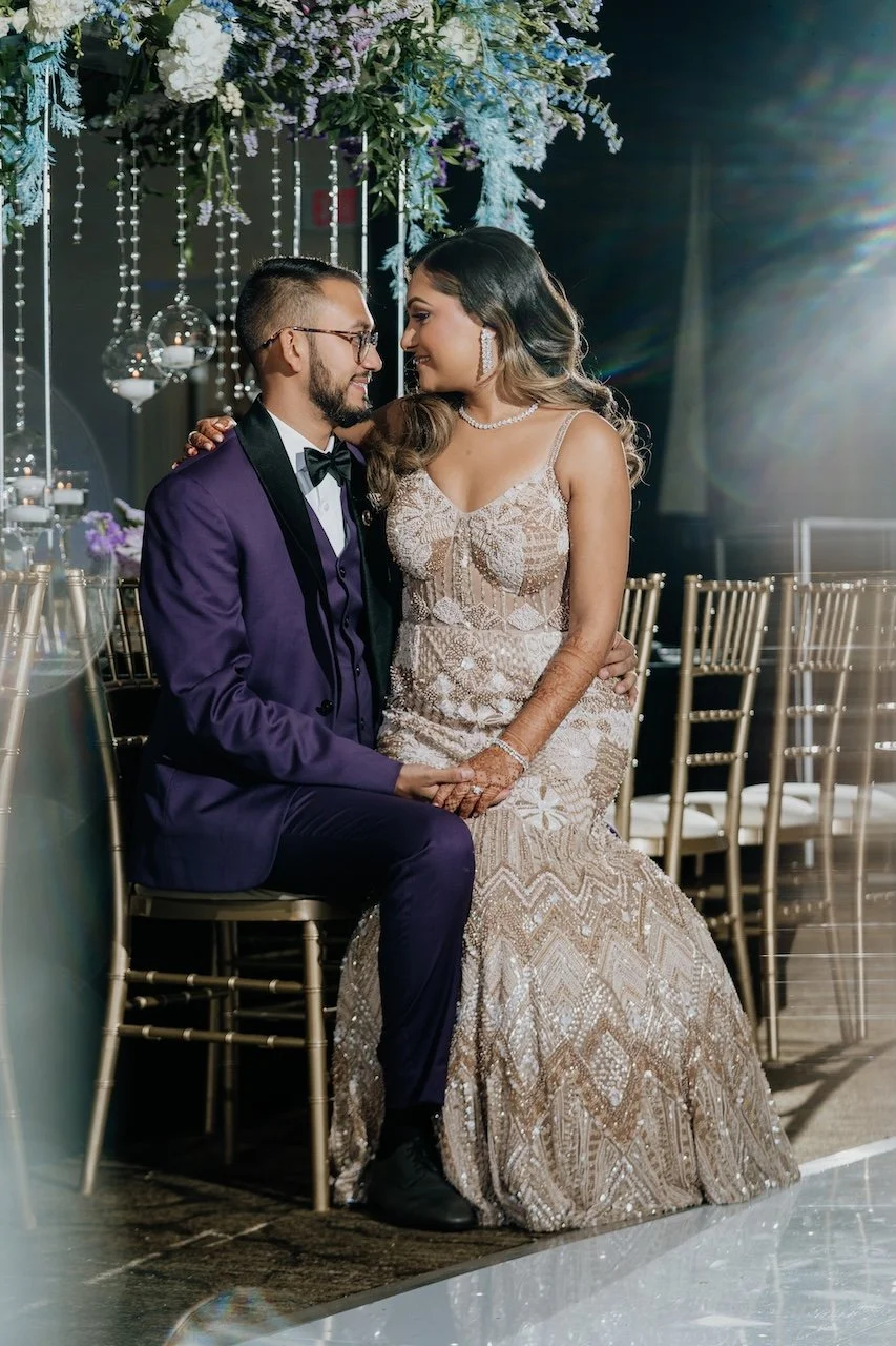 A couple dressed in formal attire sharing an intimate moment at their wedding reception, seated closely with their foreheads touching, surrounded by floral decorations and hanging ornaments.