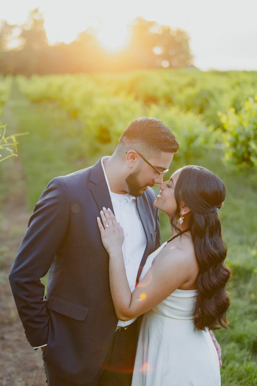 A couple in formal attire standing close together in a vineyard at sunset, smiling and touching noses.