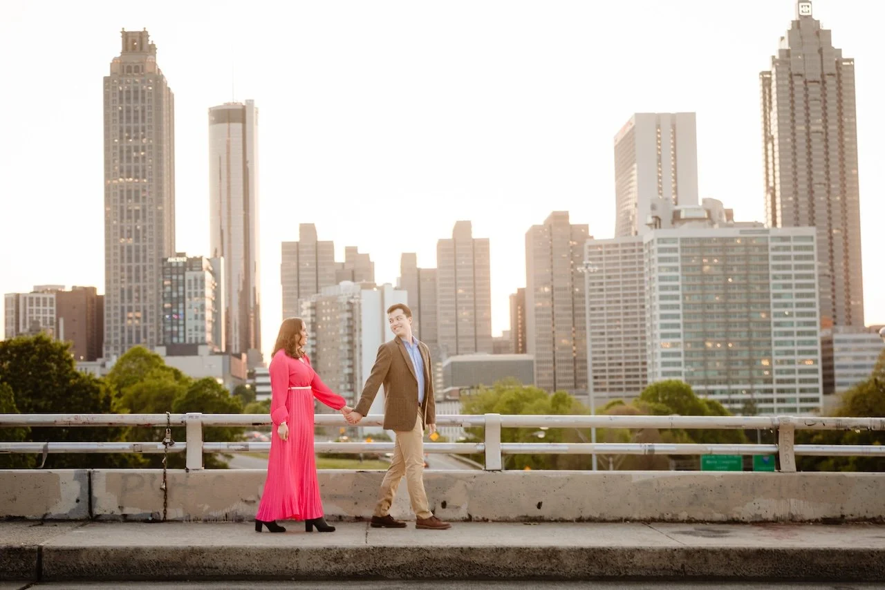 A couple holding hands on a city bridge during sunset with tall skyscrapers in the background.