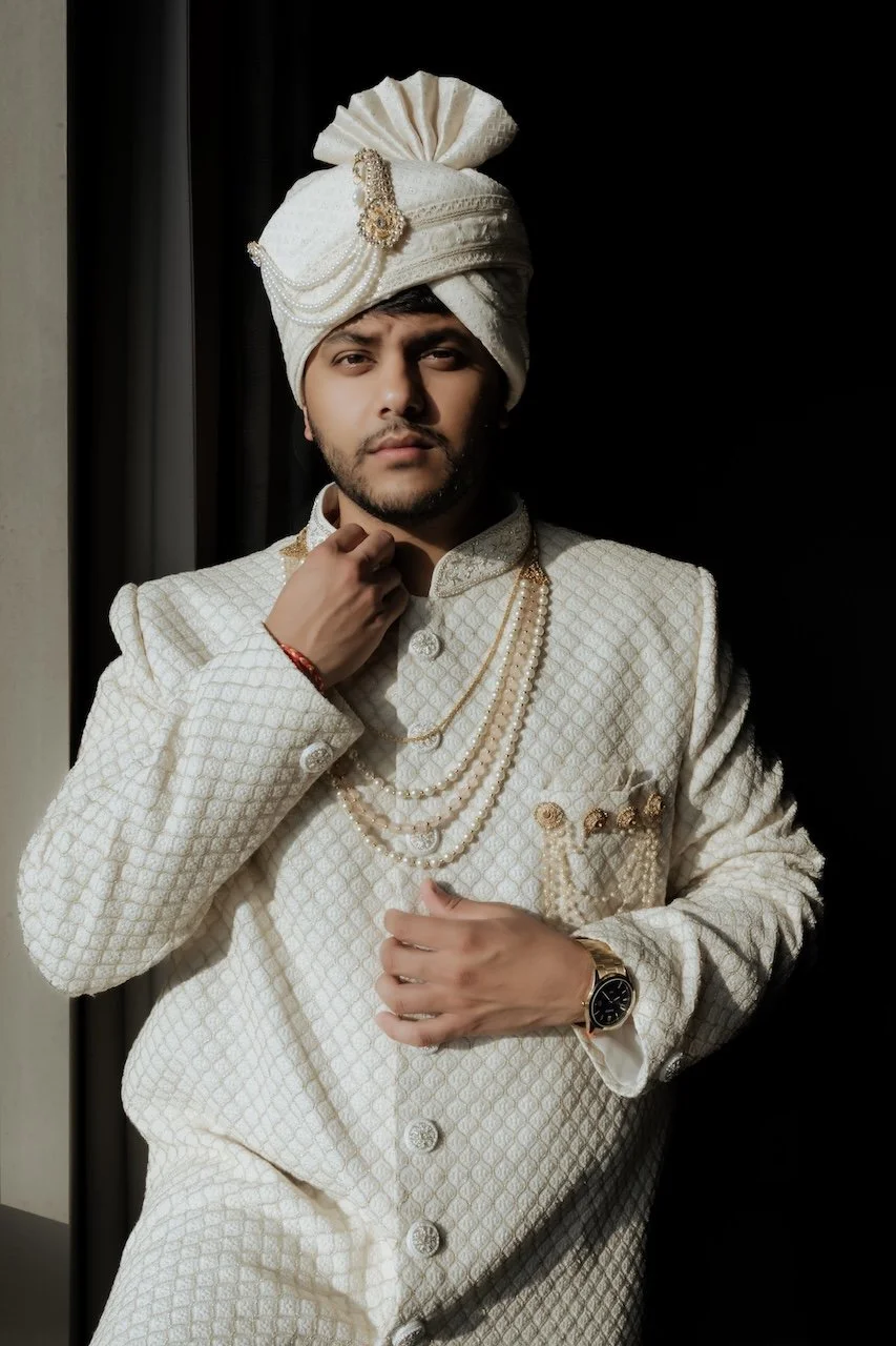 A man dressed in traditional Indian wedding attire, wearing a white sherwani, pearl necklaces, wristwatch, and a turban with embellishments, standing against a dark background.