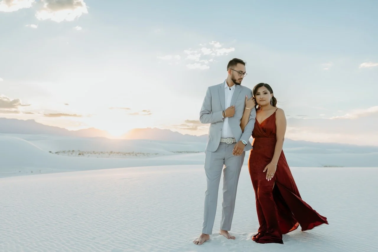 A couple standing on white sand dunes during sunset, the man in a light gray suit and the woman in a long red dress, holding hands and gazing at each other.
