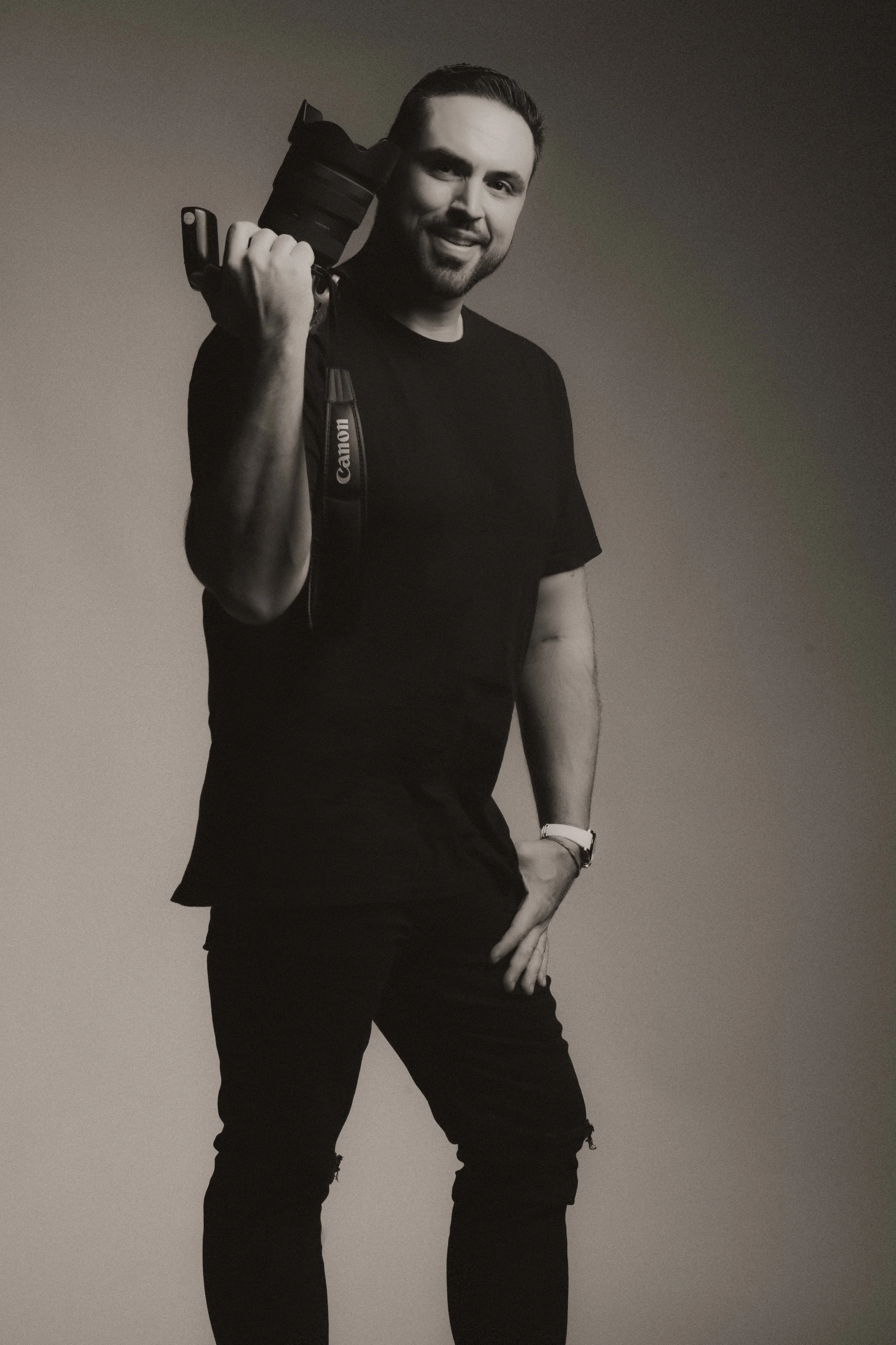 A smiling man with short hair and a beard, wearing a black t-shirt and black pants, holding a Canon camera over his shoulder, standing against a plain background.