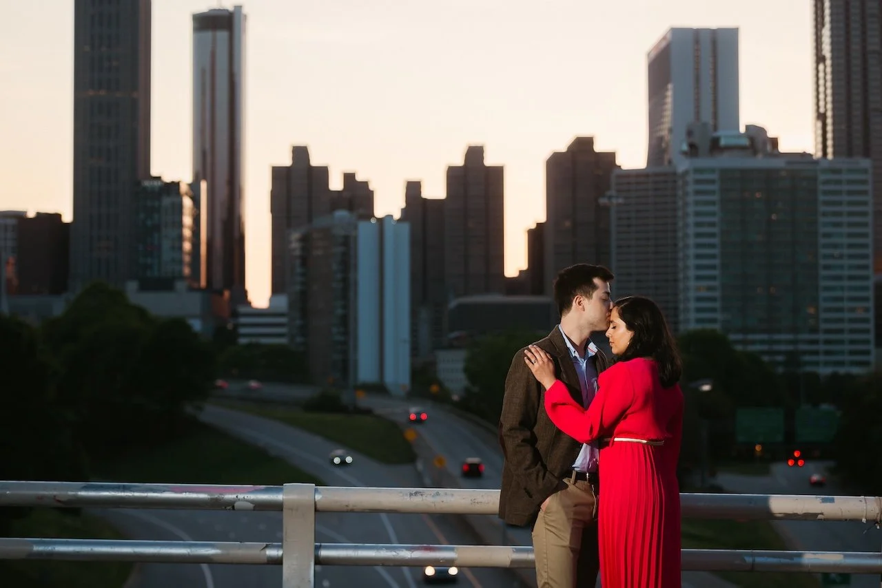 A couple standing close together on a bridge or balcony, with city skyscrapers in the background during sunset, sharing an intimate moment.