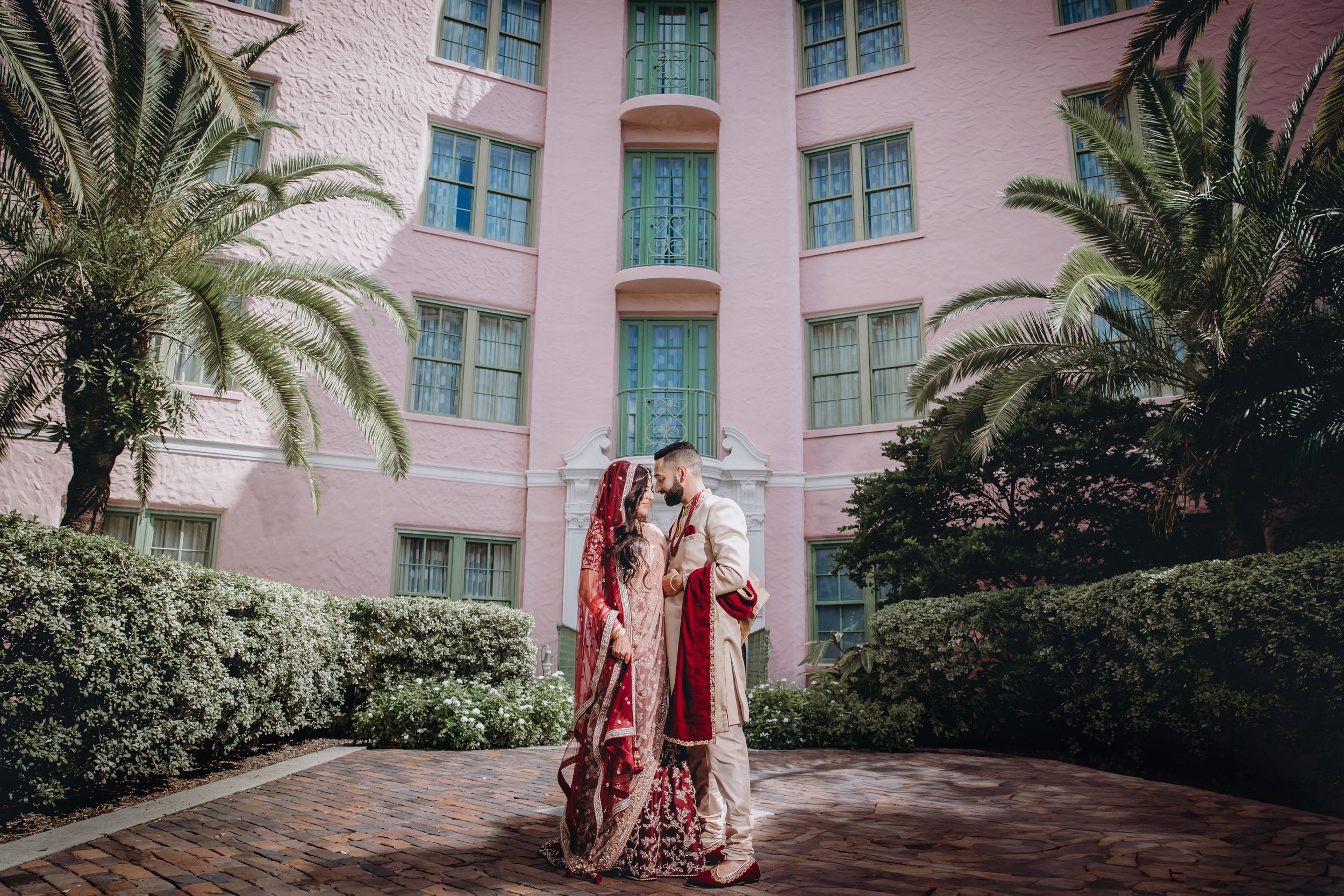 A bride and groom in traditional South Asian wedding attire stand close together in front of a pink building with green windows, holding hands and smiling at each other amidst lush greenery.