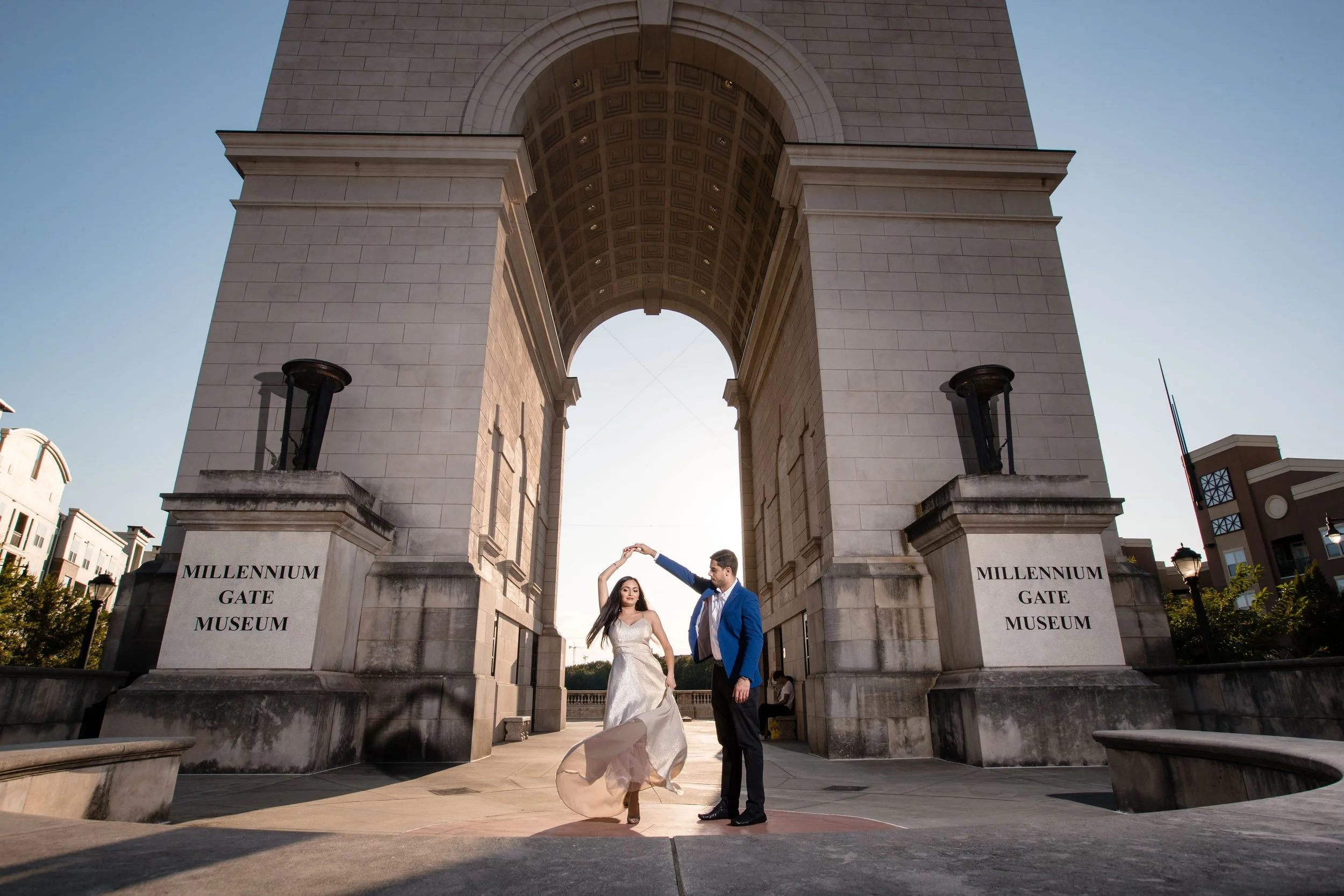 A couple dancing underneath the Millennium Gate, a large triumphal arch structure with two signs reading 'MILLENNIUM GATE MUSEUM' on either side. The woman is wearing a white dress and the man is dressed in a blue blazer and black pants, holding her 