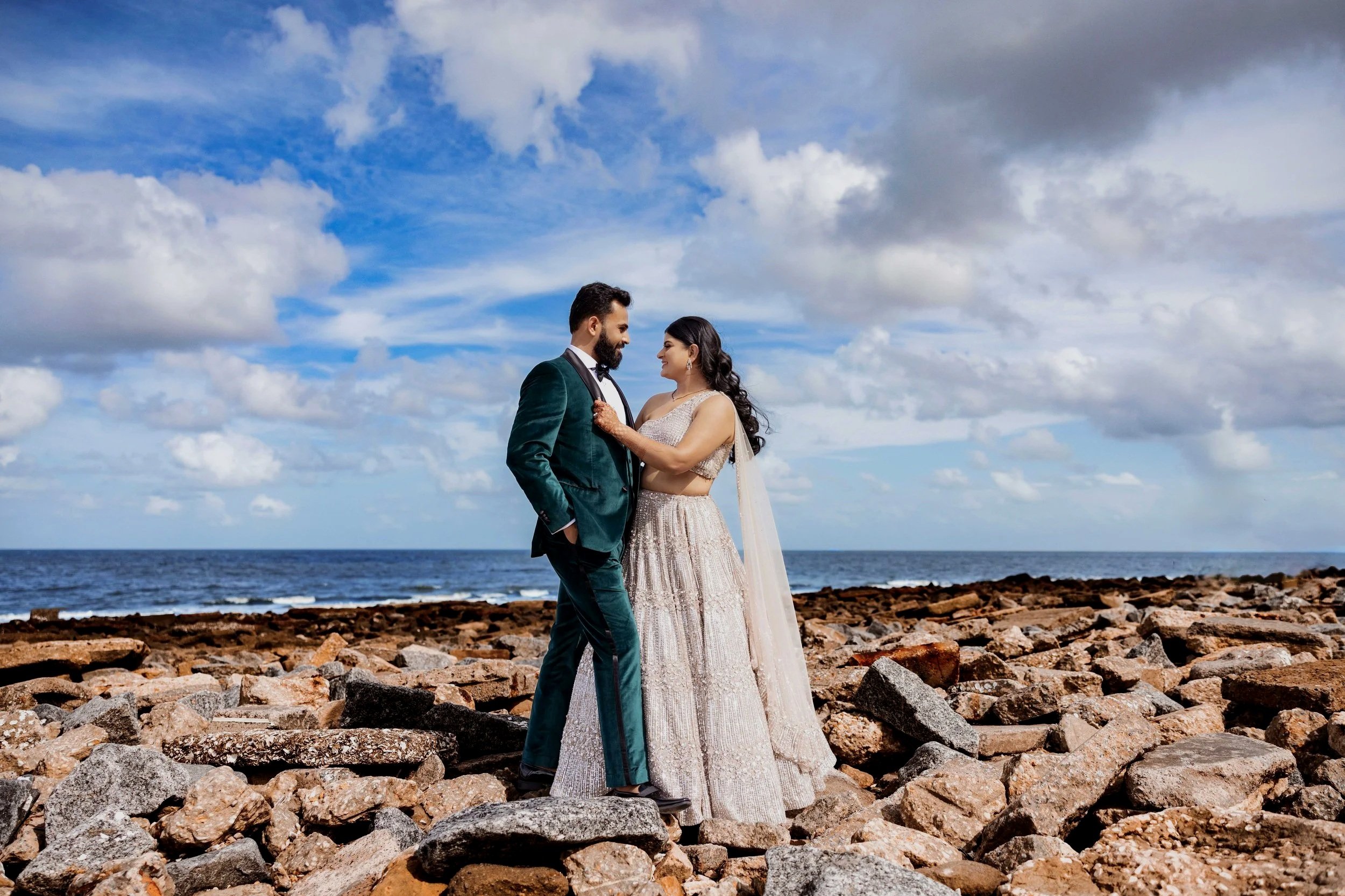 A couple dressed in wedding attire standing on rocky beach with ocean and partly cloudy sky in the background.