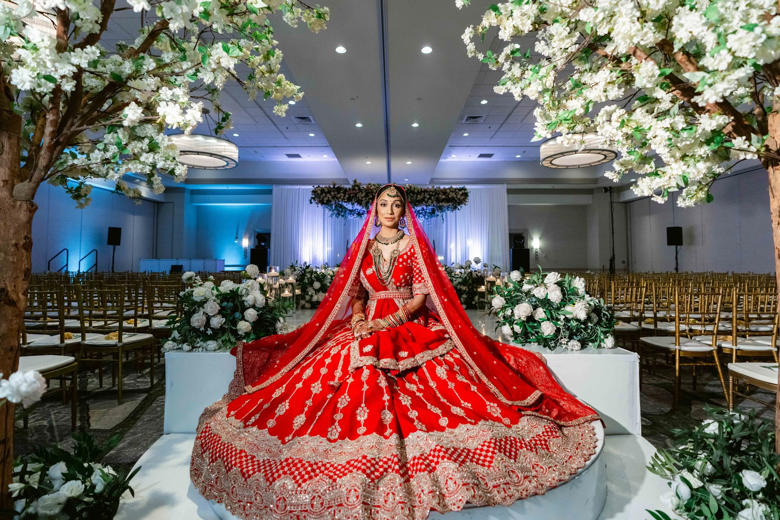 A bride in a red wedding dress with gold embroidery, sitting on a white platform in a decorated wedding hall with white flowers, trees, and rows of chairs.