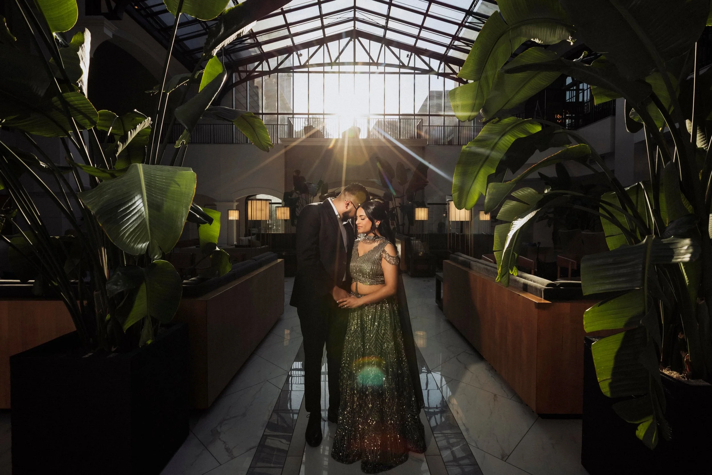 A couple dressed in formal attire, standing close and holding hands, is inside a spacious building with a glass ceiling. The sunlight is shining through the ceiling, creating a lens flare. Large green plants frame the couple on either side, and the i