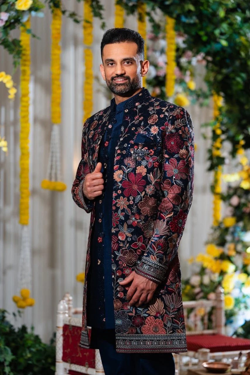 A man dressed in a dark floral embroidered sherwani, standing in front of a decorated floral backdrop with yellow flowers, for an event or celebration.