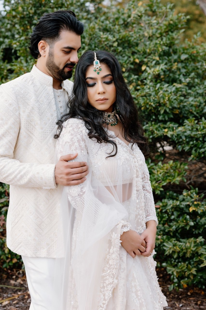 A man and woman dressed in traditional Indian wedding attire standing close together outdoors, with the man gently holding the woman's shoulder. The woman is wearing elaborate jewelry, including a maang tikka and choker, and has long, wavy hair. Thei